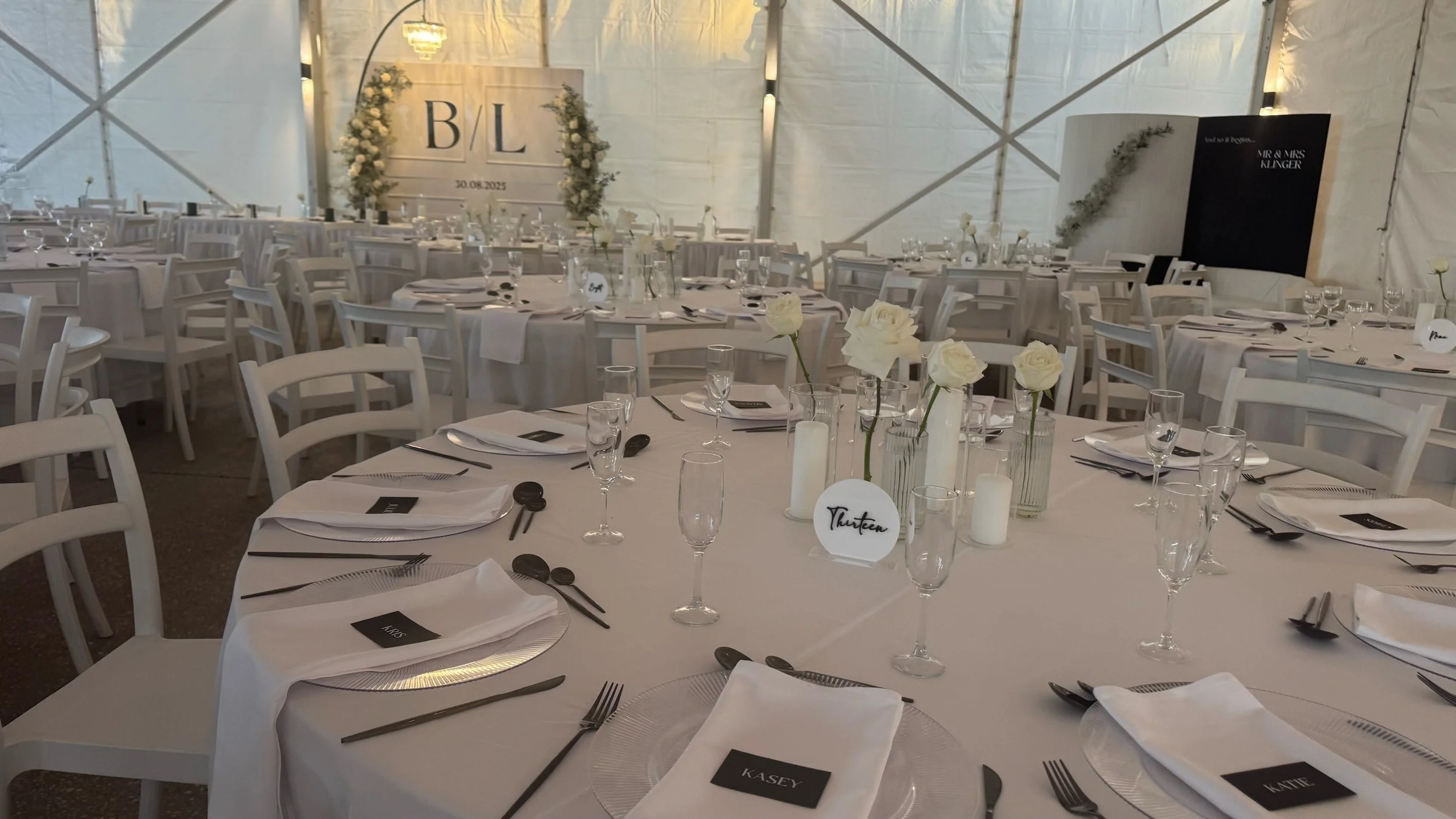 Wedding reception setup with round tables, white chairs, white tablecloths, floral centerpieces, candles, and place cards inside a tent.