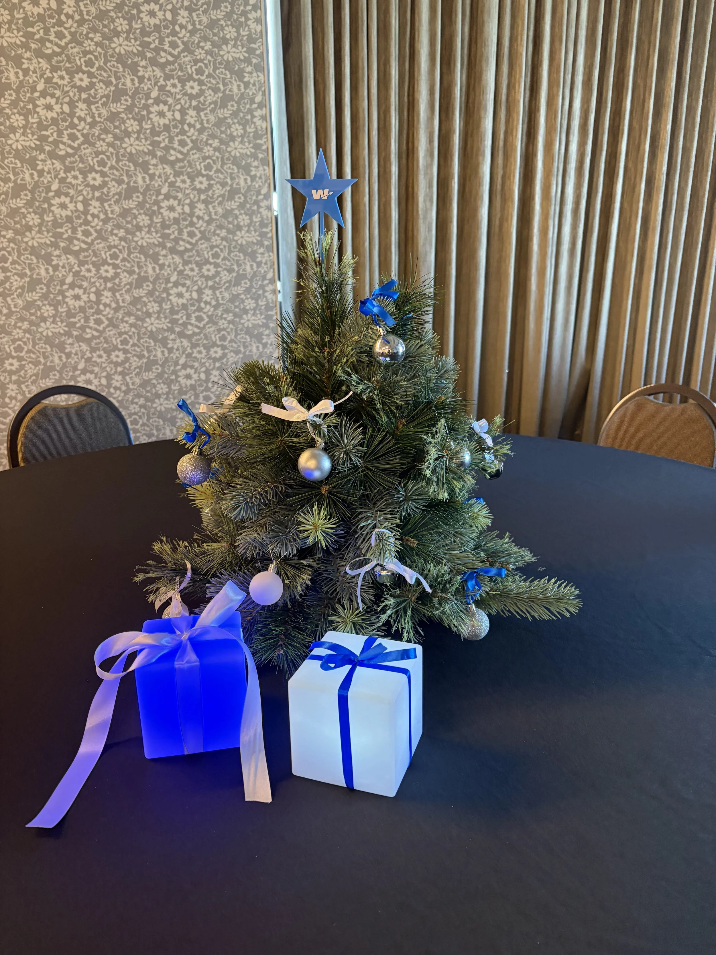 Small decorated Christmas tree on a black table with two wrapped presents in blue and white ribbons, set against a background of a wood-paneled wall and floral-patterned wallpaper.