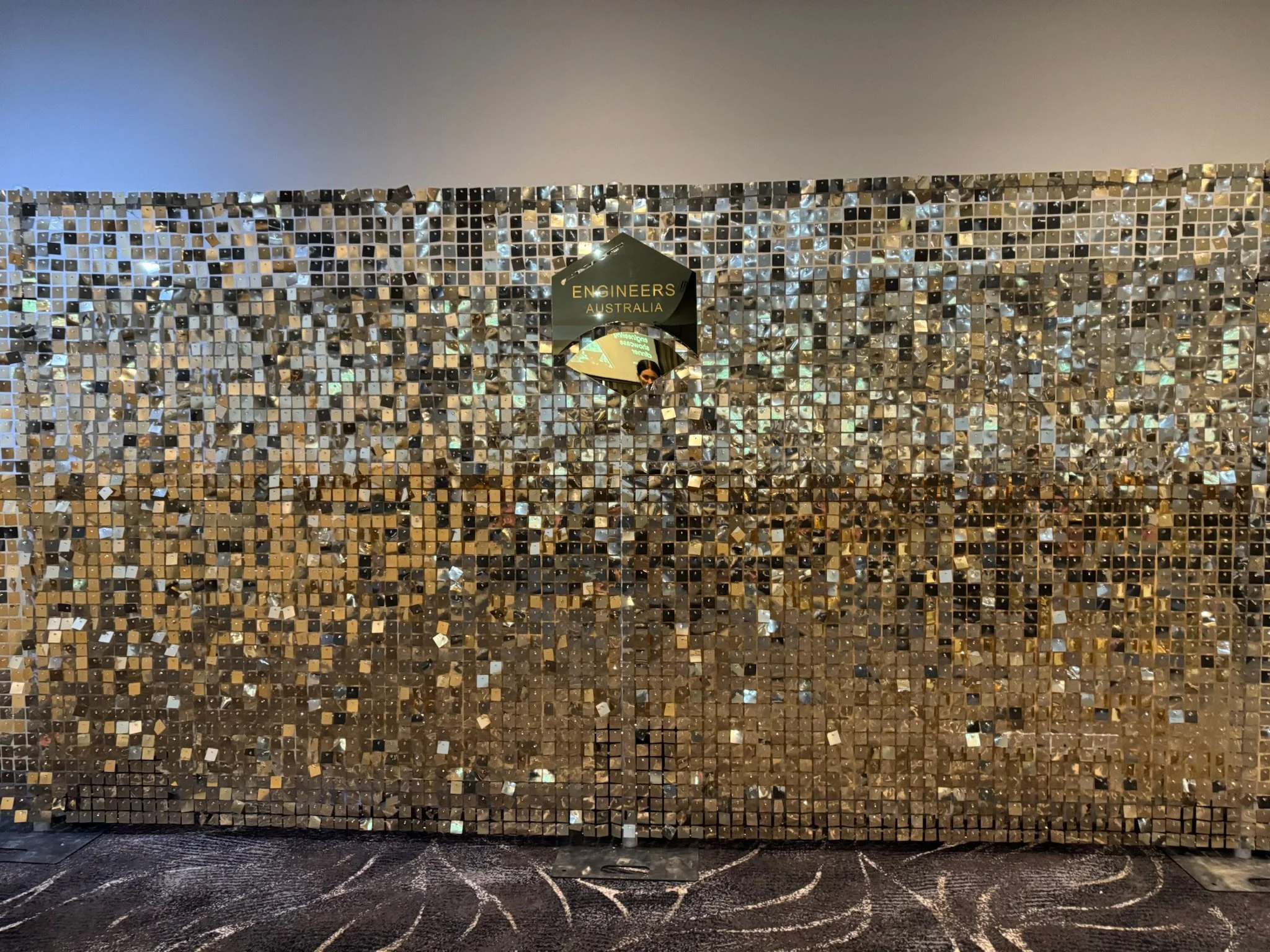 A wall covered in small, shiny, square tiles in gold and silver colors, with a sign reading 'Engineers Australia' and a partial reflection of a person taking the photo.