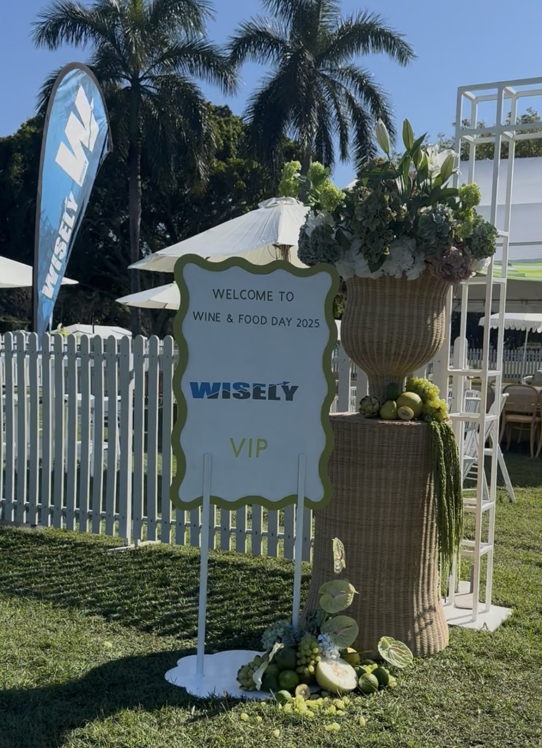 Sign welcoming guests to Wine & Food Day 2025 at Wisely with a decorative woven basket filled with flowers, surrounded by green produce and foliage, set outdoors with white umbrellas, white fencing, and palm trees in the background.