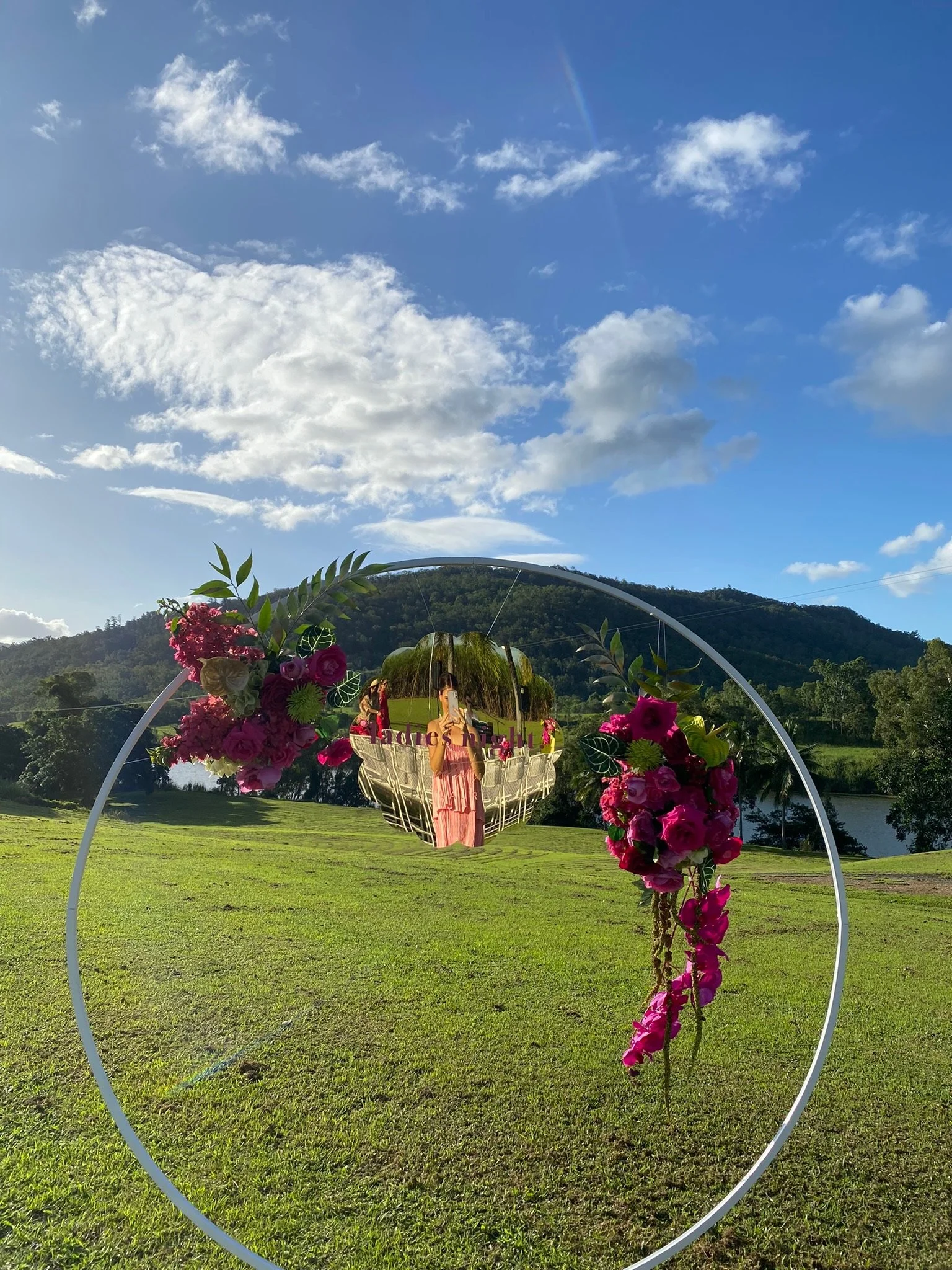 A circular mirror with pink and green floral decorations on a grassy field, reflecting a woman in a pink dress taking a photo, with mountains, trees, and a blue sky with clouds in the background.