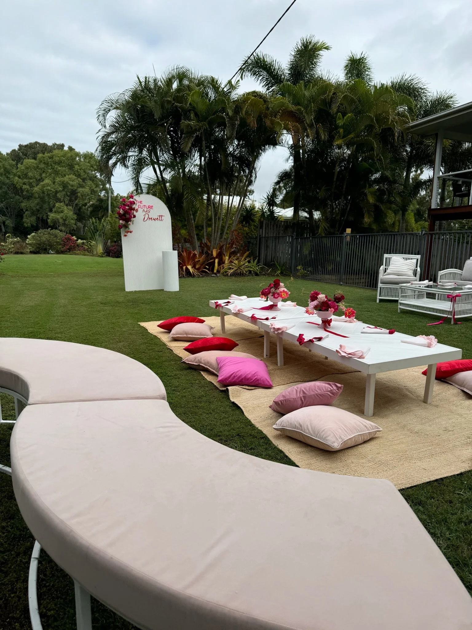 Outdoor garden setup with low white table, pink and red cushions, flower arrangements, and a white board with pink flowers that reads 'The Future is Female' and 'Drewett'.