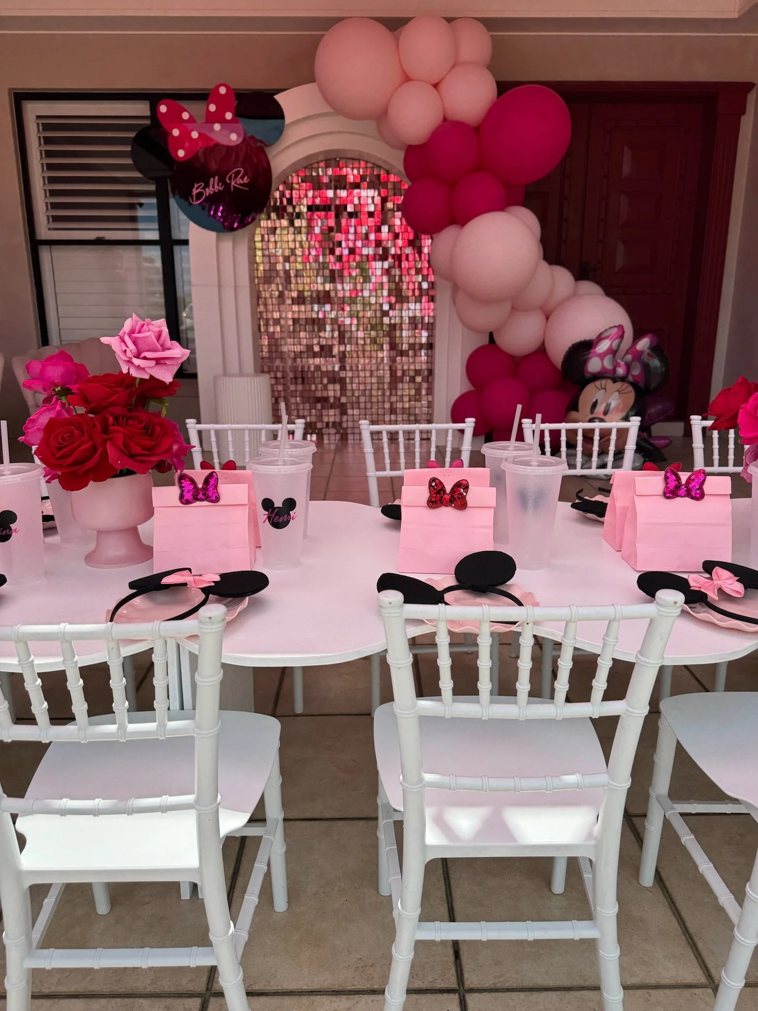 Party table decorated with pink and black Minnie Mouse theme. There are pink roses in a white vase, pink gift bags with glittery bows, Minnie Mouse ear headbands, and drinks. In the background, pink and white balloons form an arch, with a mirror and 