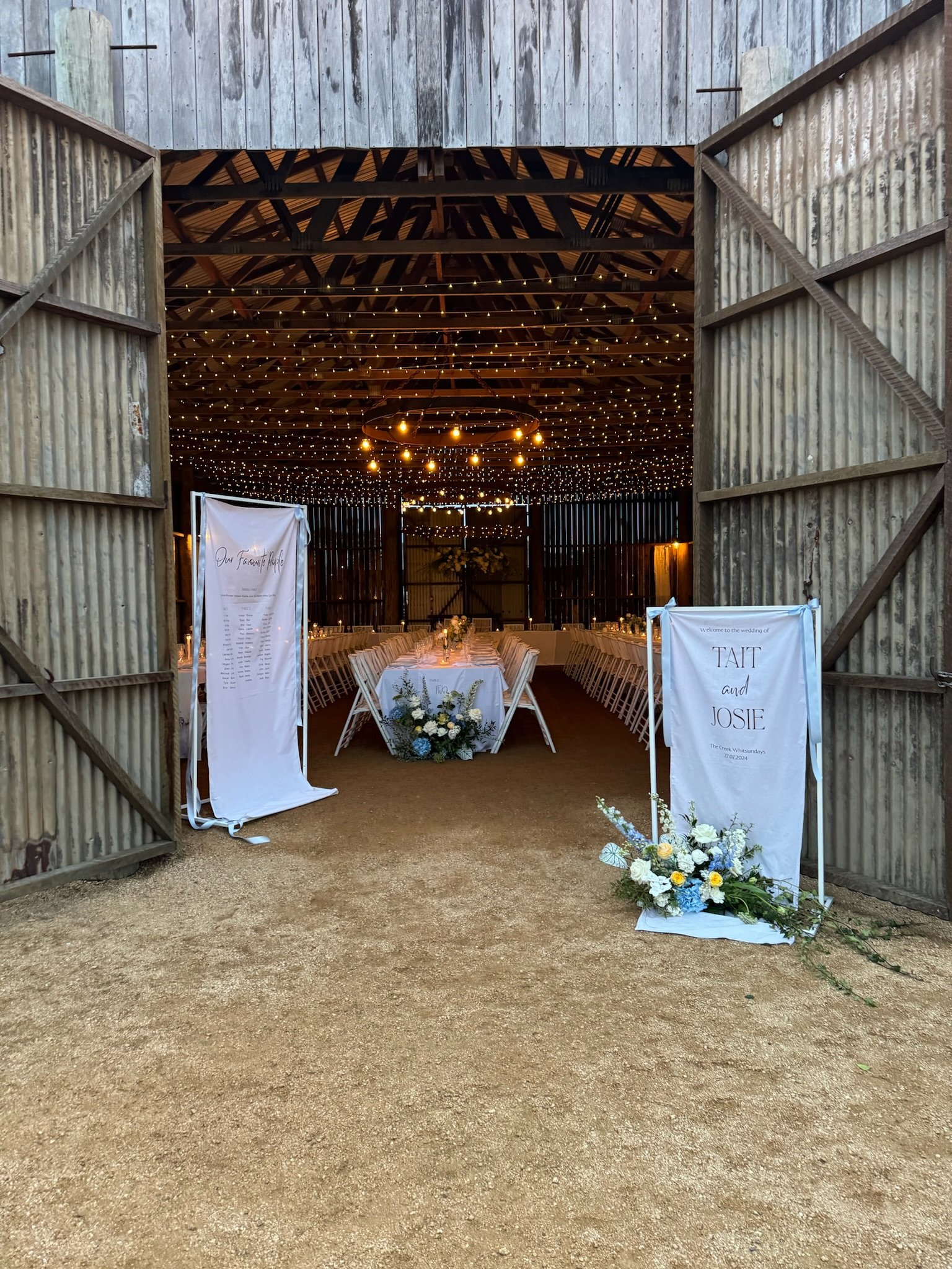 Inside a rustic barn decorated for a wedding reception with string lights hanging from the ceiling, a long table with flowers, and welcoming signs at the entrance.