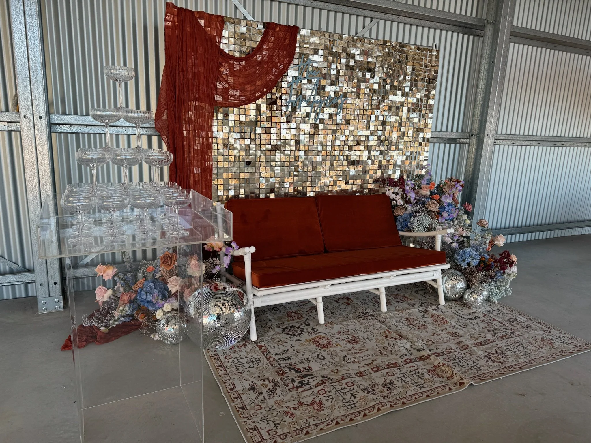 Decorative setup with a small vintage white bench with red cushions, a floral arrangement, a glass tower holding champagne glasses, and a backdrop featuring a mosaic mirror wall and red fabric draped over it, with disco balls on the floor and a patte
