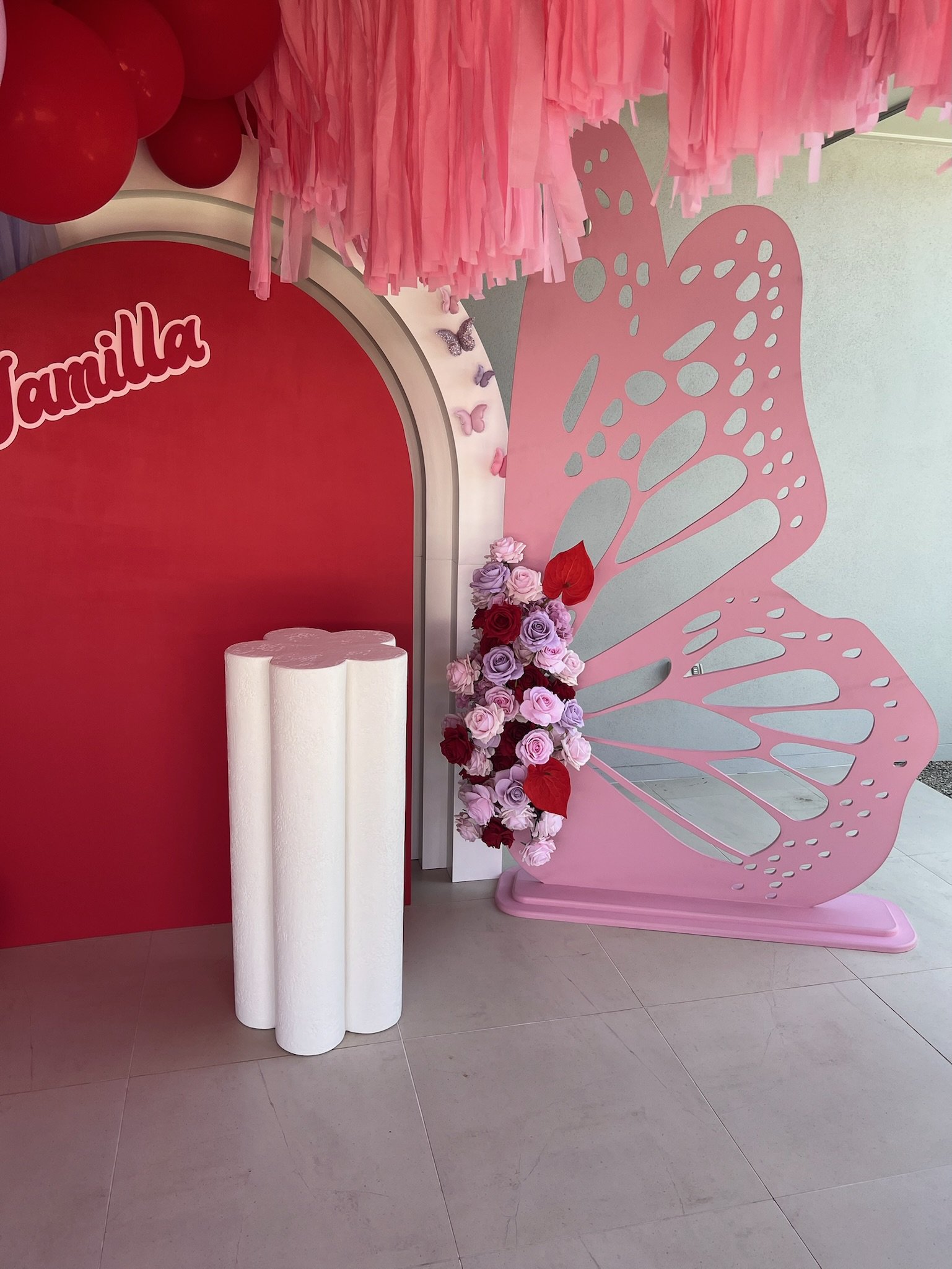 Decorative display featuring a pink butterfly-shaped stand, with a floral arrangement of pink and red roses, a pink fringe canopy, and a red backdrop with the partial word 'vanilla' on it.