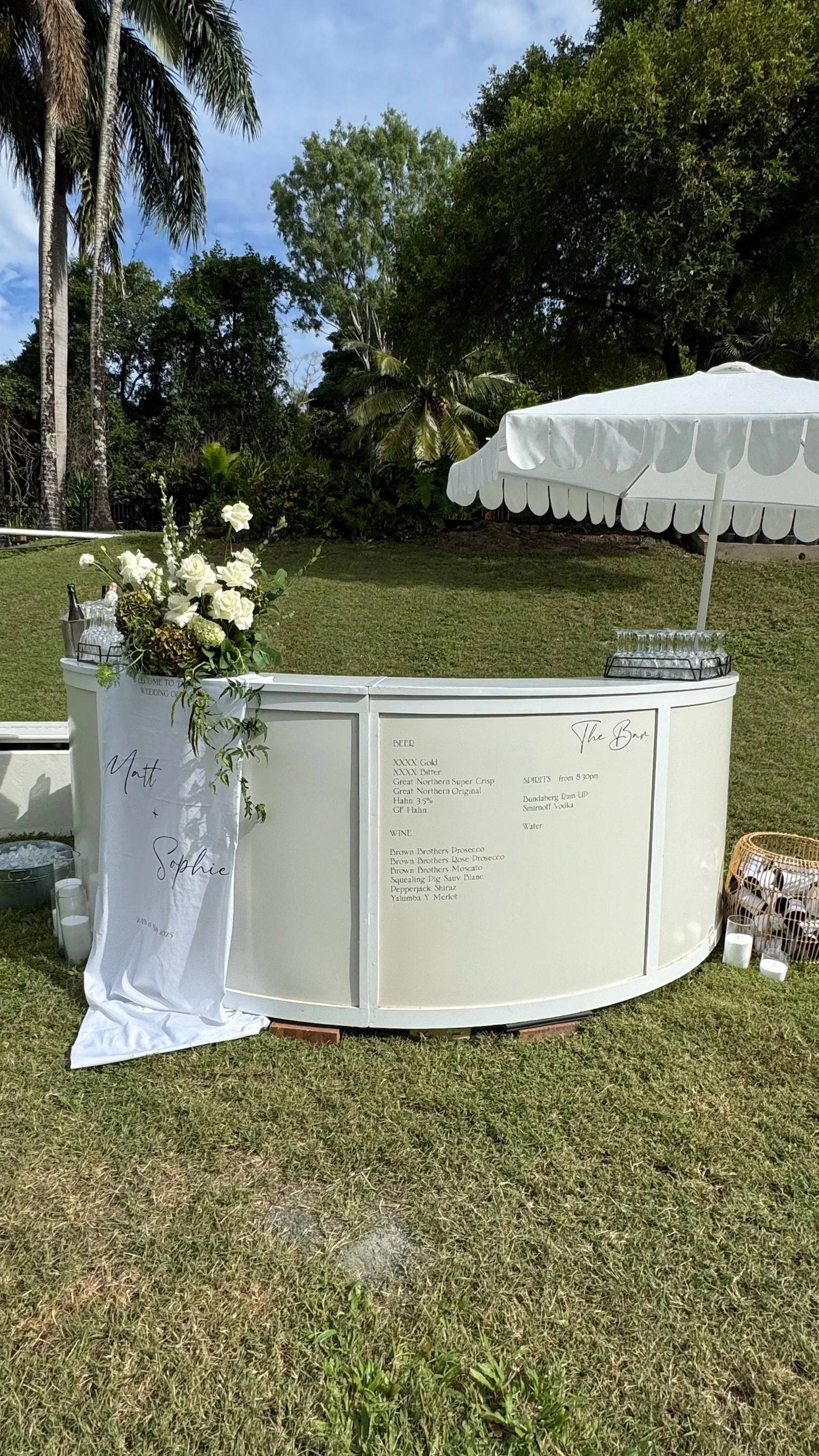 A white curved bar with a white umbrella for shade, decorated with a floral arrangement and a paper on the front with names and event details, set in a grassy outdoor area with trees in the background.