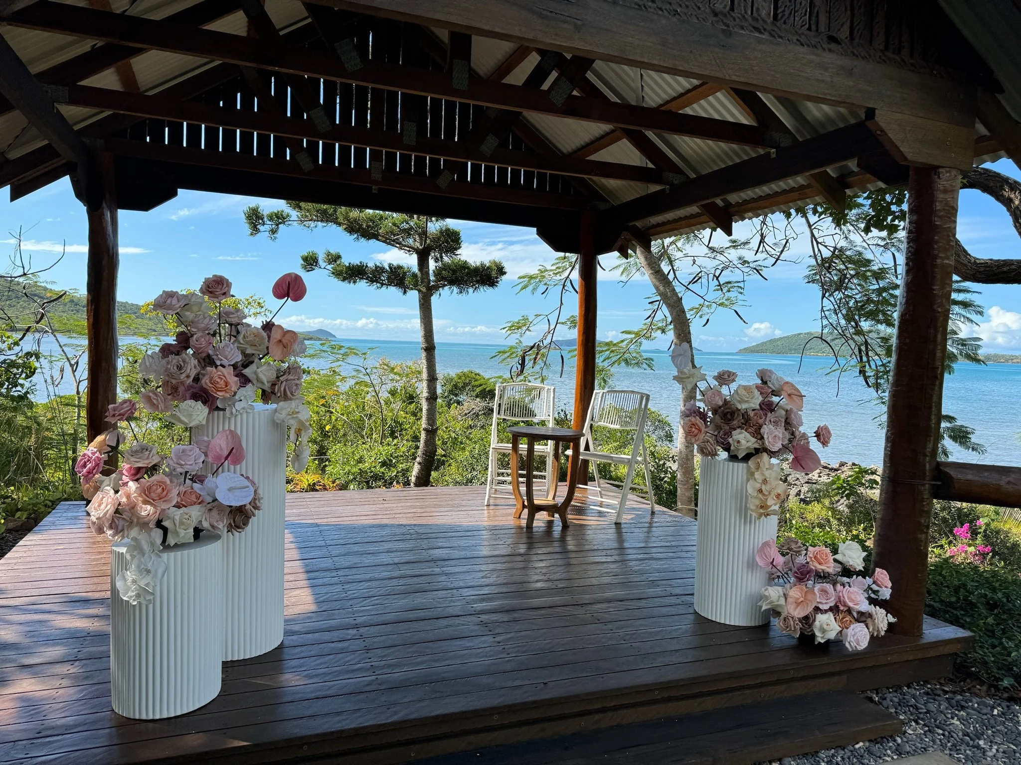 A scenic outdoor stage with flower arrangements, two white chairs, and a small table, overlooking a body of water with small islands and trees surrounding the area.