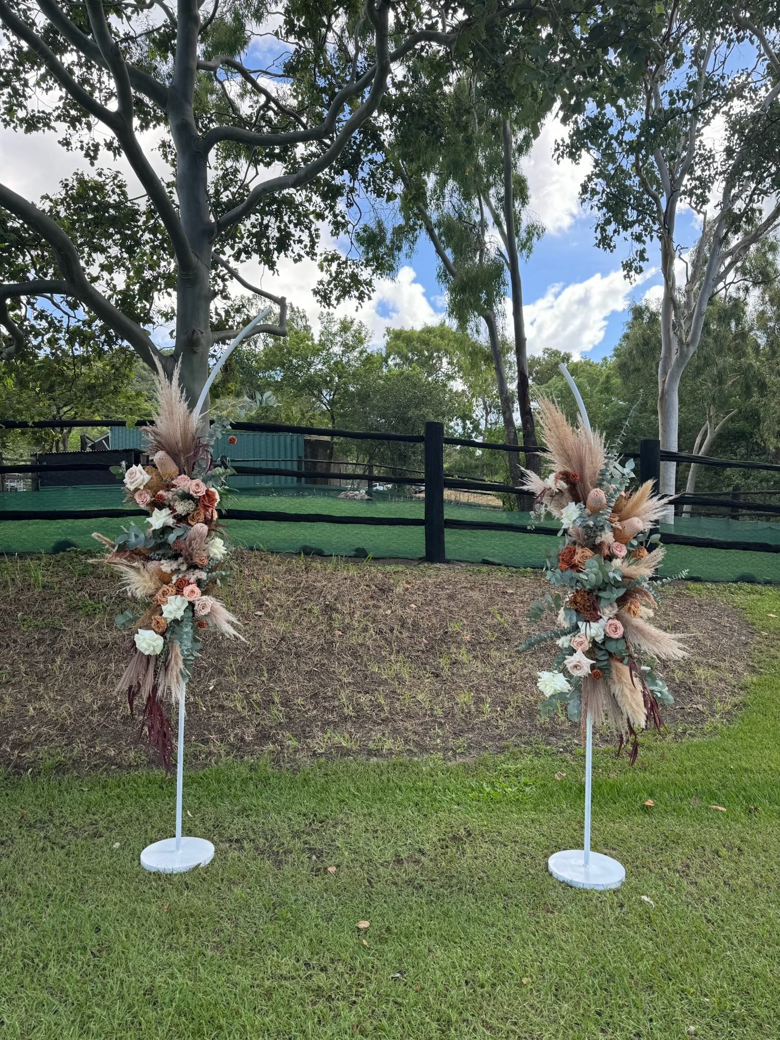 Two decorative floral arrangements on white stands outdoors on grass, with trees, a black fence, and a cloudy sky in the background.