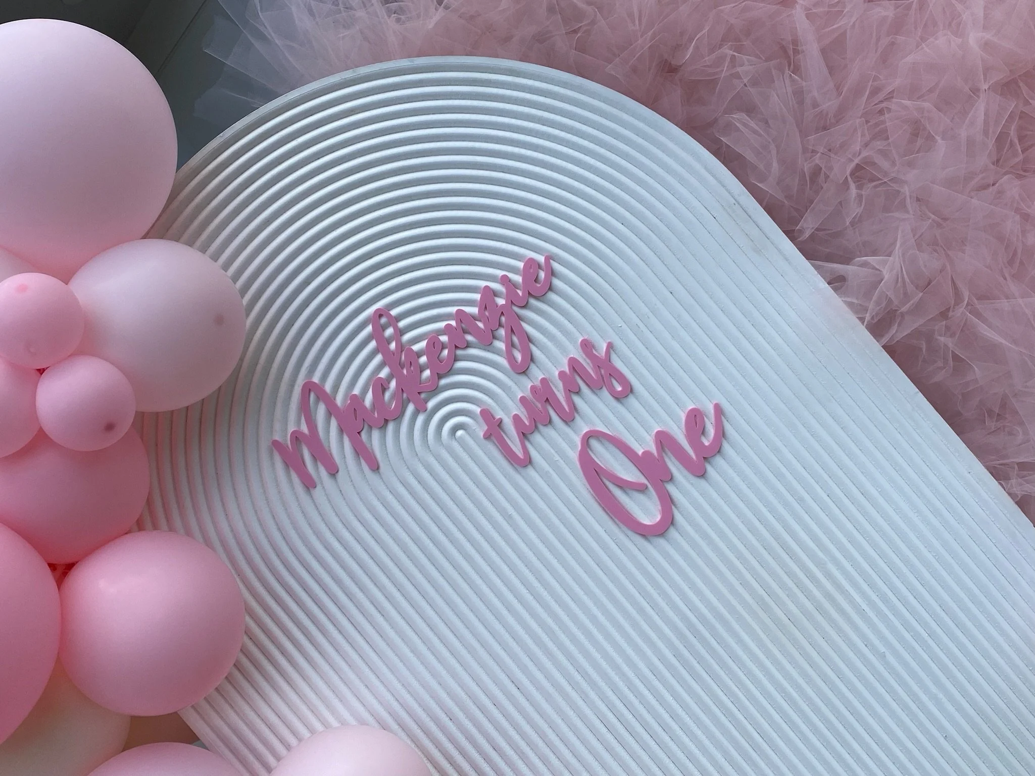 Decorative display with pink balloons arranged around a white textured board with pink cursive text that reads "Mackenzie turning One," and soft pink tulle fabric in the background.