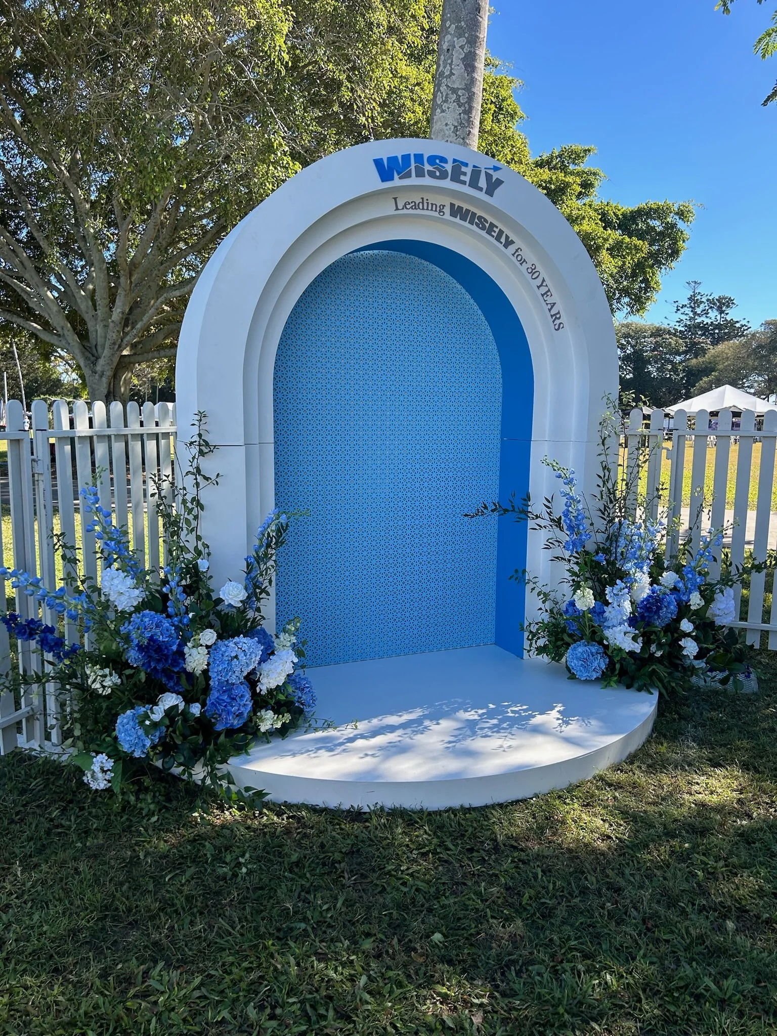A decorative outdoor display with a white and blue arch reading 'WISELY Leading WISELY for 30 YEARS,' decorated with blue and white flowers at its base, placed in a grassy area with trees and a clear blue sky in the background.