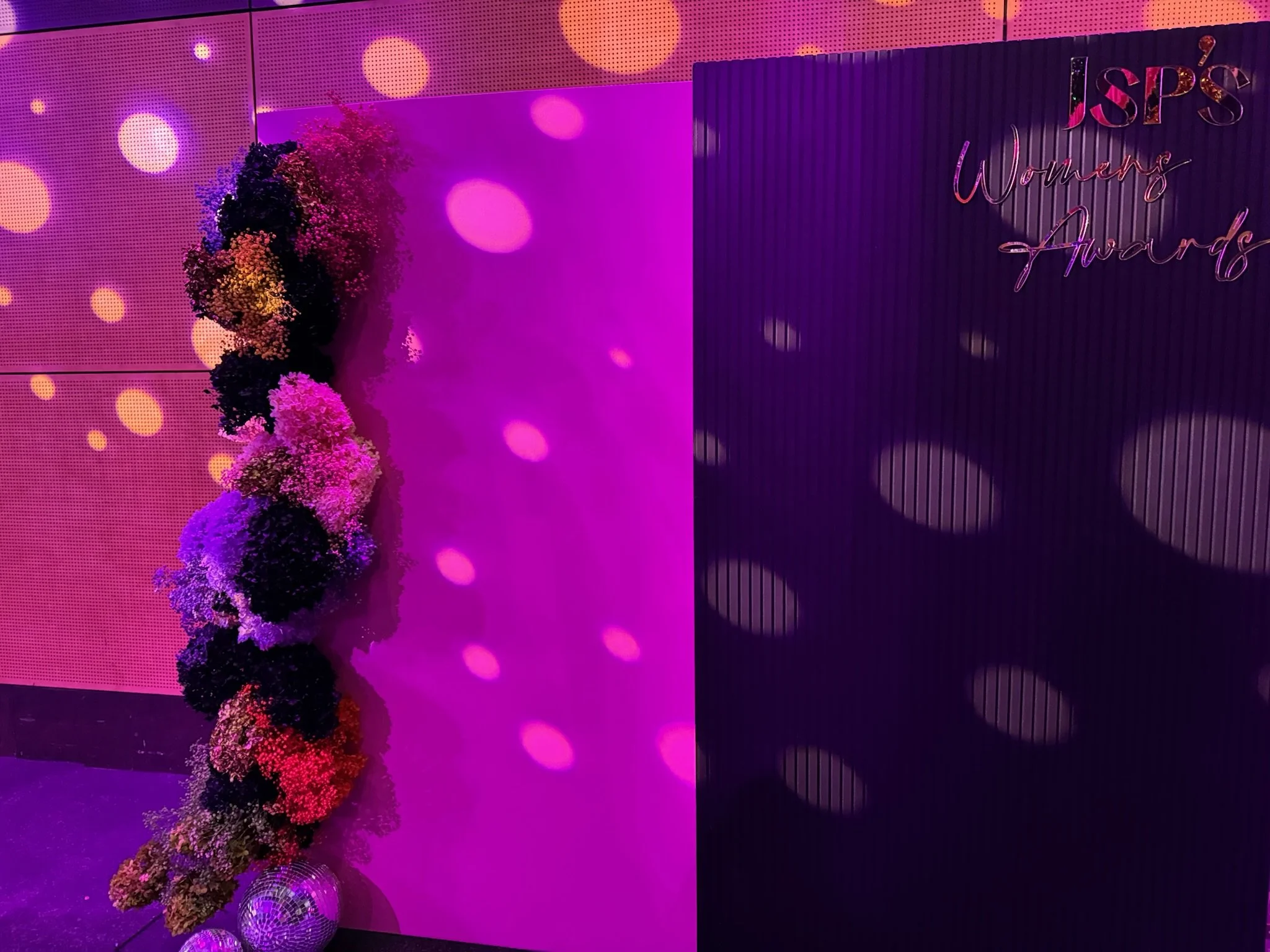 Decorative floral arrangement with colorful blooms on a side, with purple and pink lighting, and a sign that reads 'JSP's Women's Awards' on a dark background.