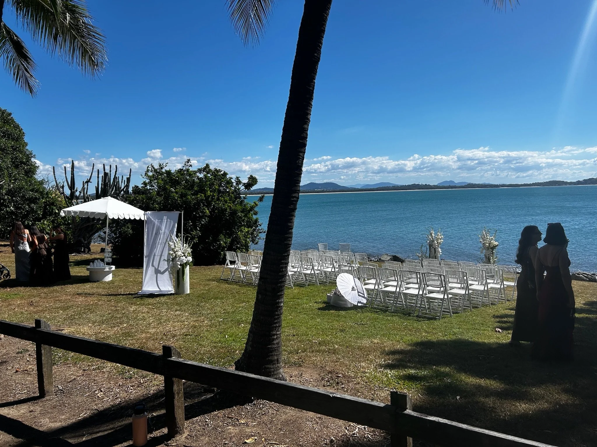 Setup for an outdoor wedding ceremony by the water with white chairs, floral arrangements, and a white arch on a grassy area under a blue sky.