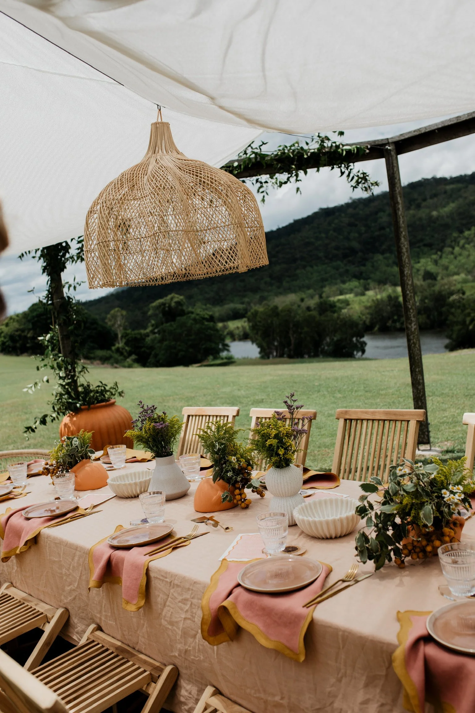 Outdoor dining table set under a canopy, decorated with vases of flowers, bowls, glasses, and plates, with a scenic view of green hills and river in the background.