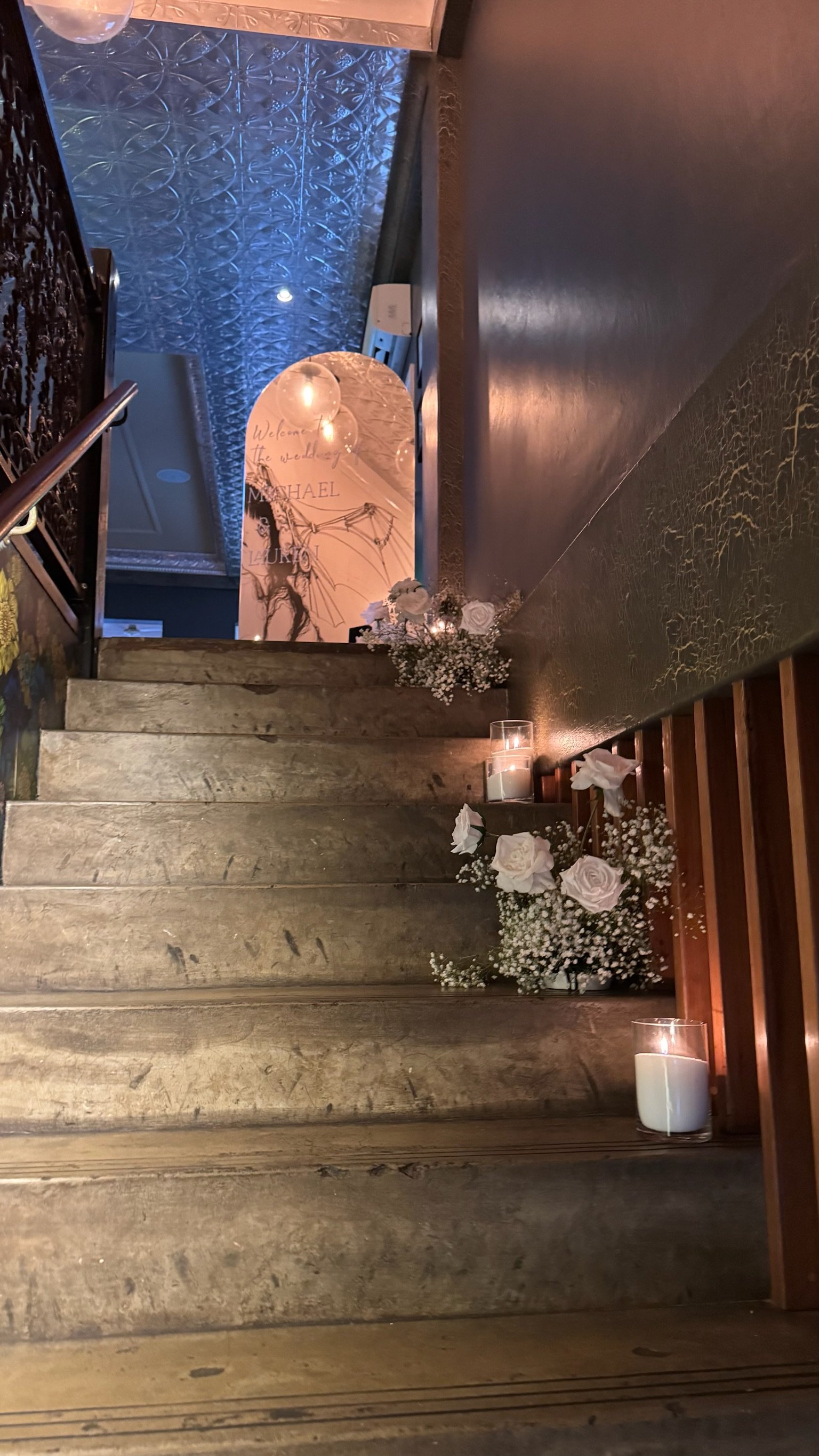 Indoor staircase decorated with white roses, baby's breath flowers, and candles for a wedding. At the top of the stairs, a mirror with a handwritten message and floral arrangements is visible.