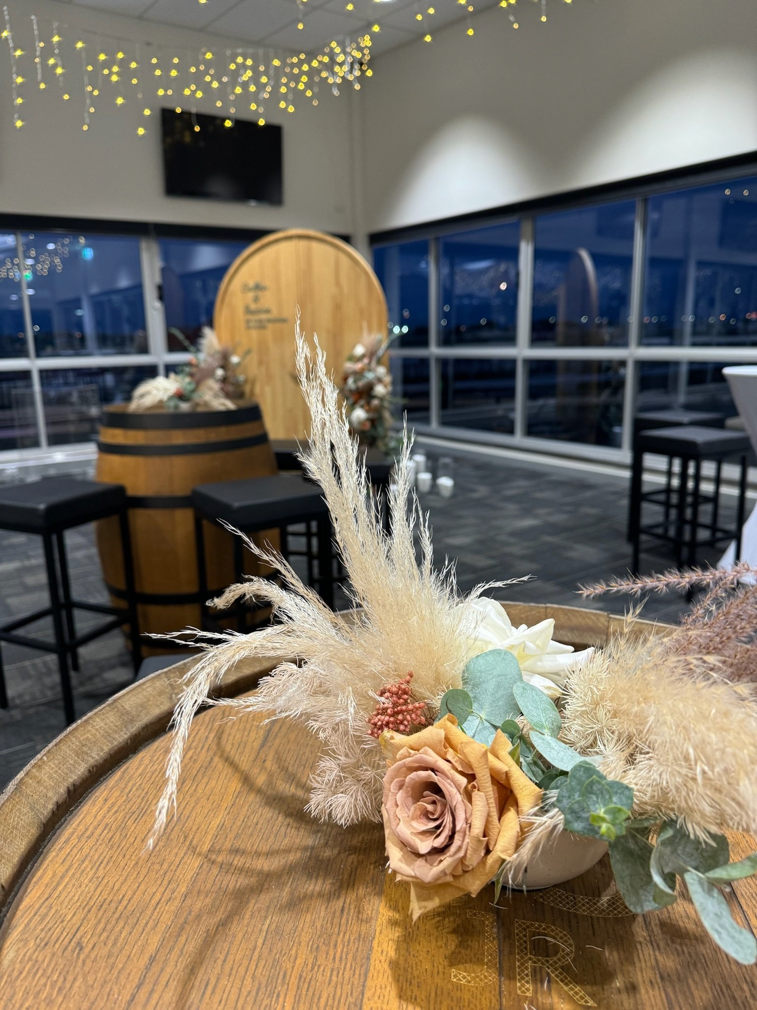 Decorative floral arrangement with a cream rose, dried pampas grass, eucalyptus leaves in a white bowl on a wooden table. In the background is a rustic bar setup with barstools, floral arrangements, and string lights, with large windows showing a nig