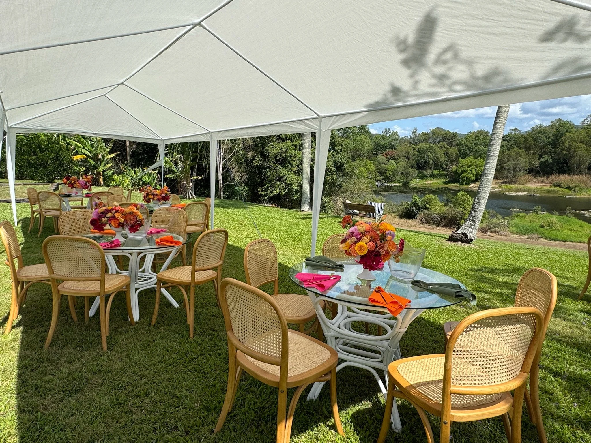 Outdoor event setup with round tables under a white canopy tent on green grass, decorated with colorful floral centerpieces, pink and orange napkins, and surrounded by wooden chairs, overlooking a river, trees, and blue sky.