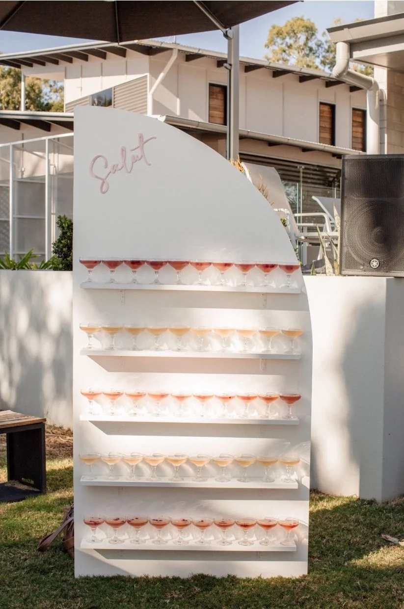 A white display stand with multiple shelves holding glasses with drinks. The stand has the word 'Salut' written in pink cursive at the top left corner. The background features a modern house and outdoor furniture.