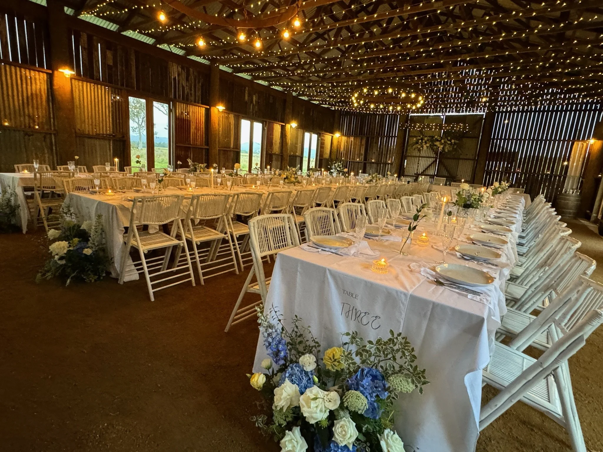 Wedding reception setup inside a barn with long tables, white chairs, floral arrangements, and string lights hanging from the wooden ceiling.