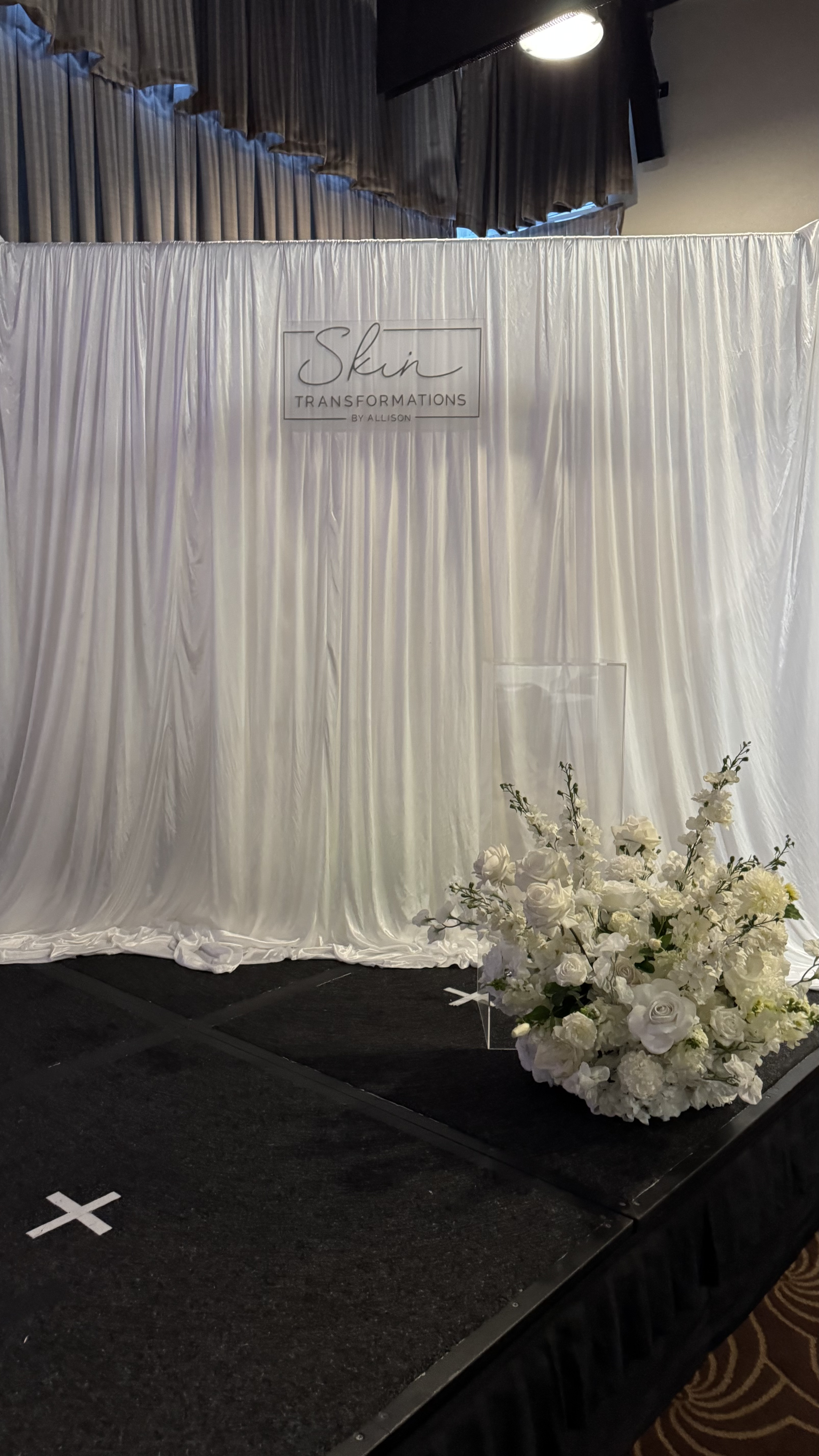 A stage with a white curtain backdrop, decorated with a floral arrangement of white roses and greenery; sign reads 'Skin Transformations by Allison'.
