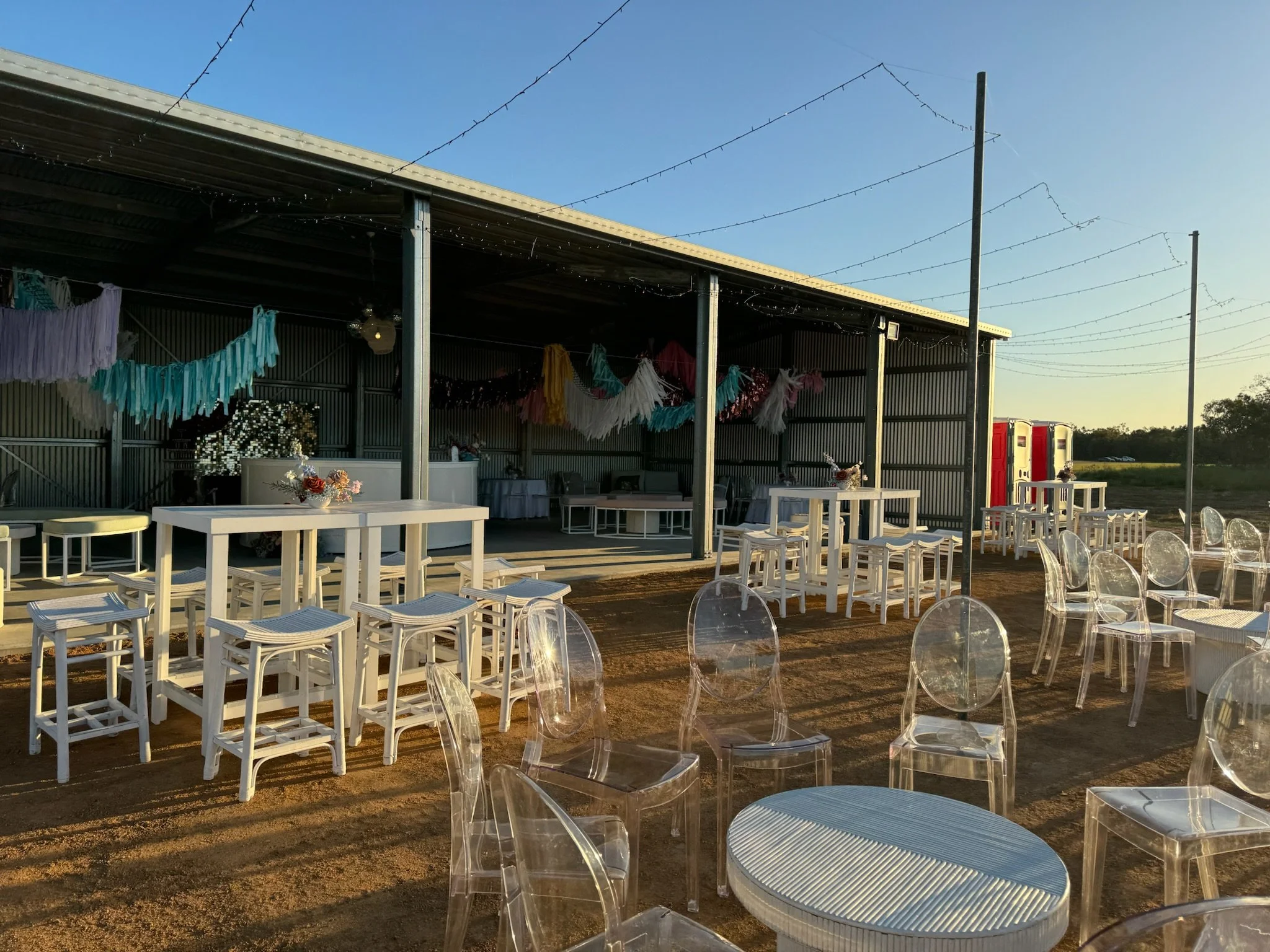 Outdoor party setup with white tables, clear chairs, colorful tassel decorations, and a metal shed in the background during sunset.