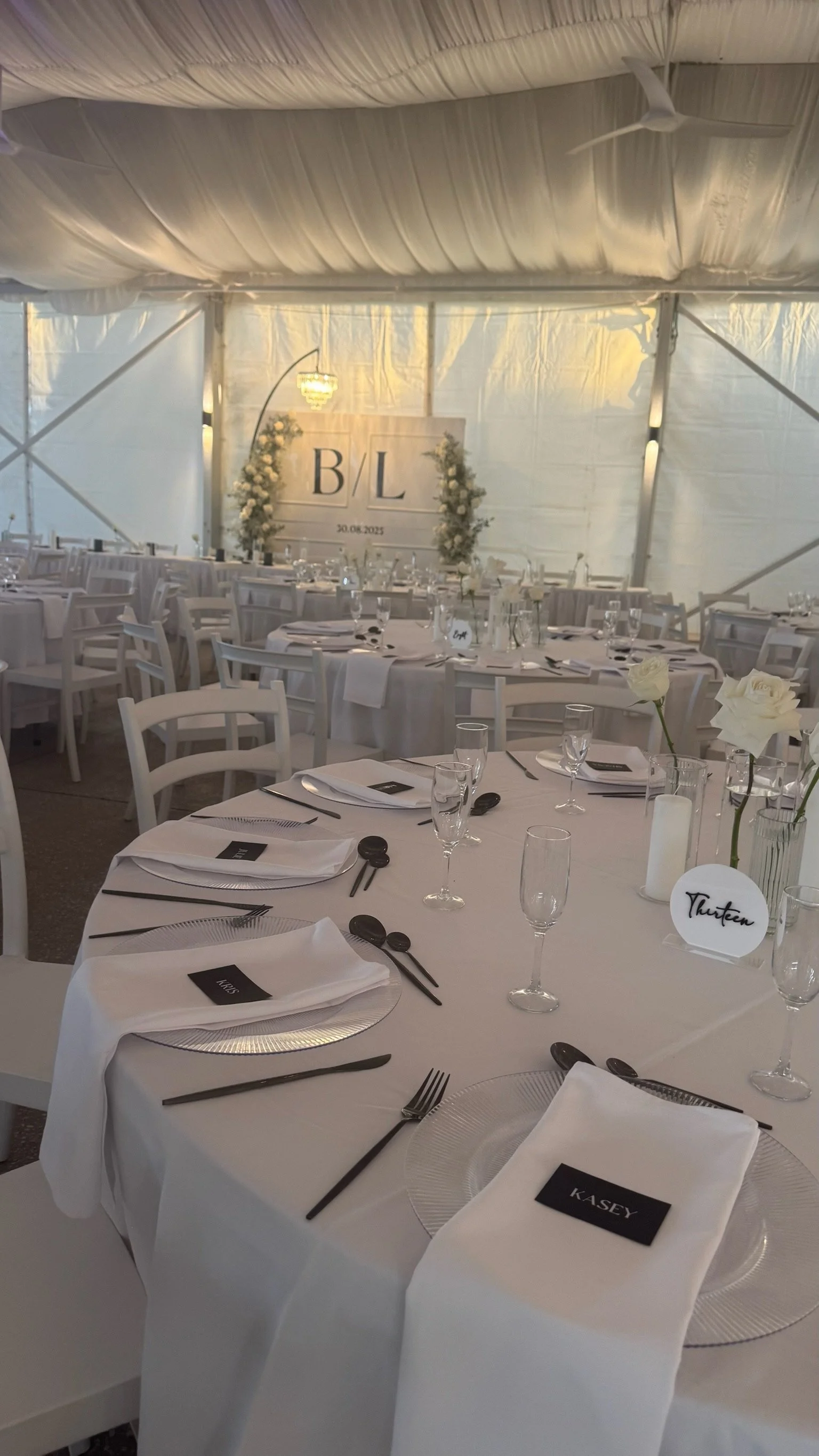 Elegant wedding reception setup inside a decorated tent with white tablecloths, silver chargers, black utensils, and white chairs. Centerpieces include white roses and candles. A large sign with 'B/L' and the date 30.08.2023 is visible in the backgro
