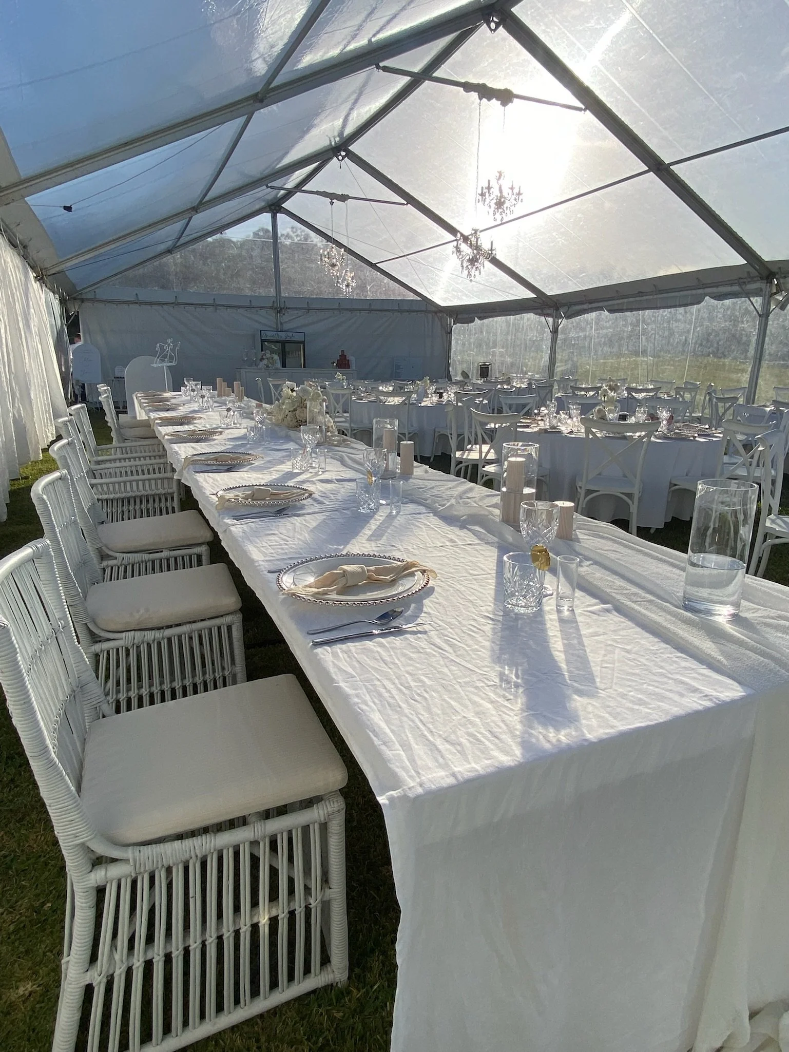 Elegant wedding reception tent with long white tablecloth-covered table set with plates, glasses, and napkins, surrounded by white chairs with cushions, and decorated with chandeliers and candles.