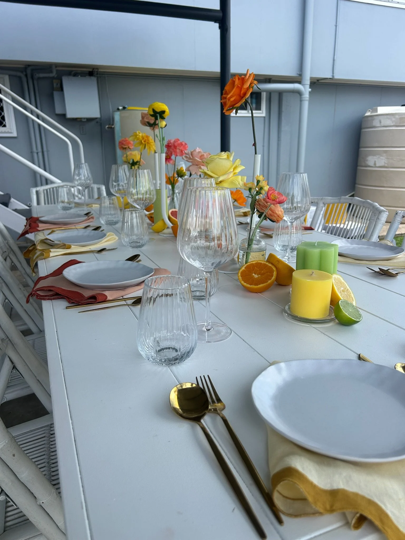 A decorated outdoor dining table set with floral centerpieces, candles, glassware, and plates with napkins.