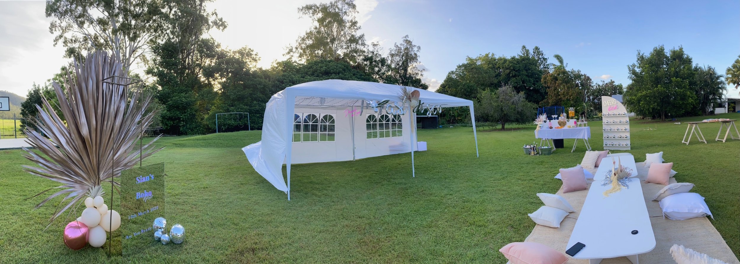 Decorative outdoor setup with a white tent, a table with snacks and drinks, a sign, and seating with cushions on a grassy field, with trees and blue sky in the background.