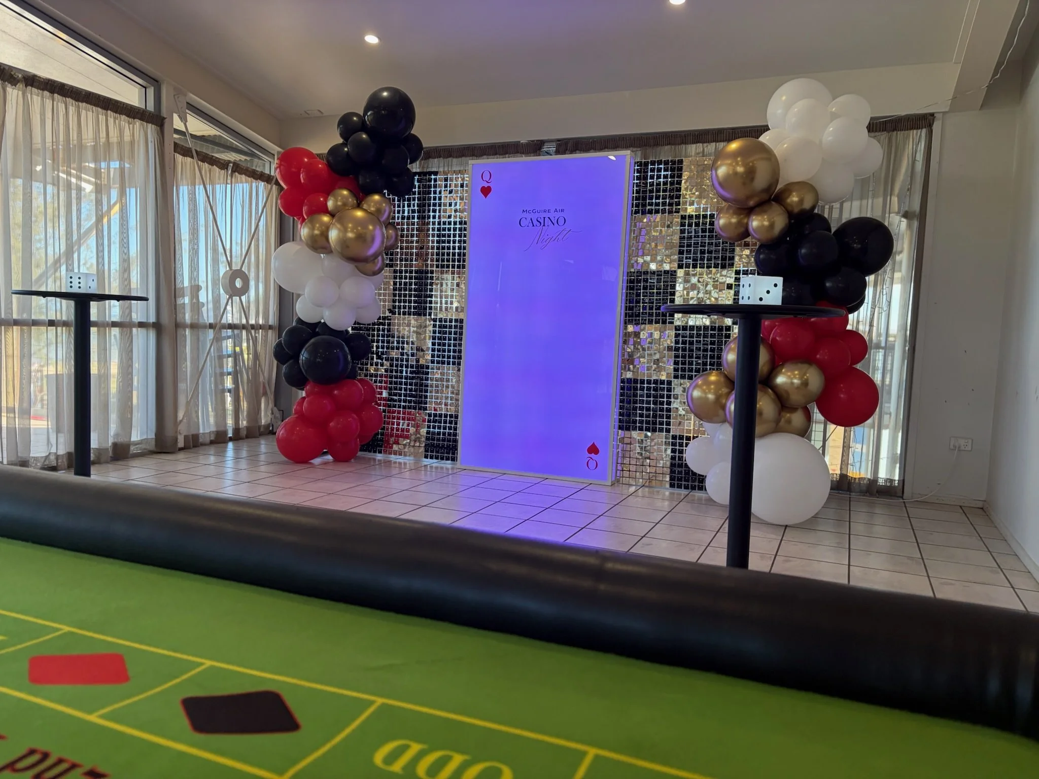 Decorated gaming or casino event area with balloon arches featuring black, gold, red, and white balloons, a large vertical screen displaying 'Casino Night,' and a T-shaped gaming table in the foreground.