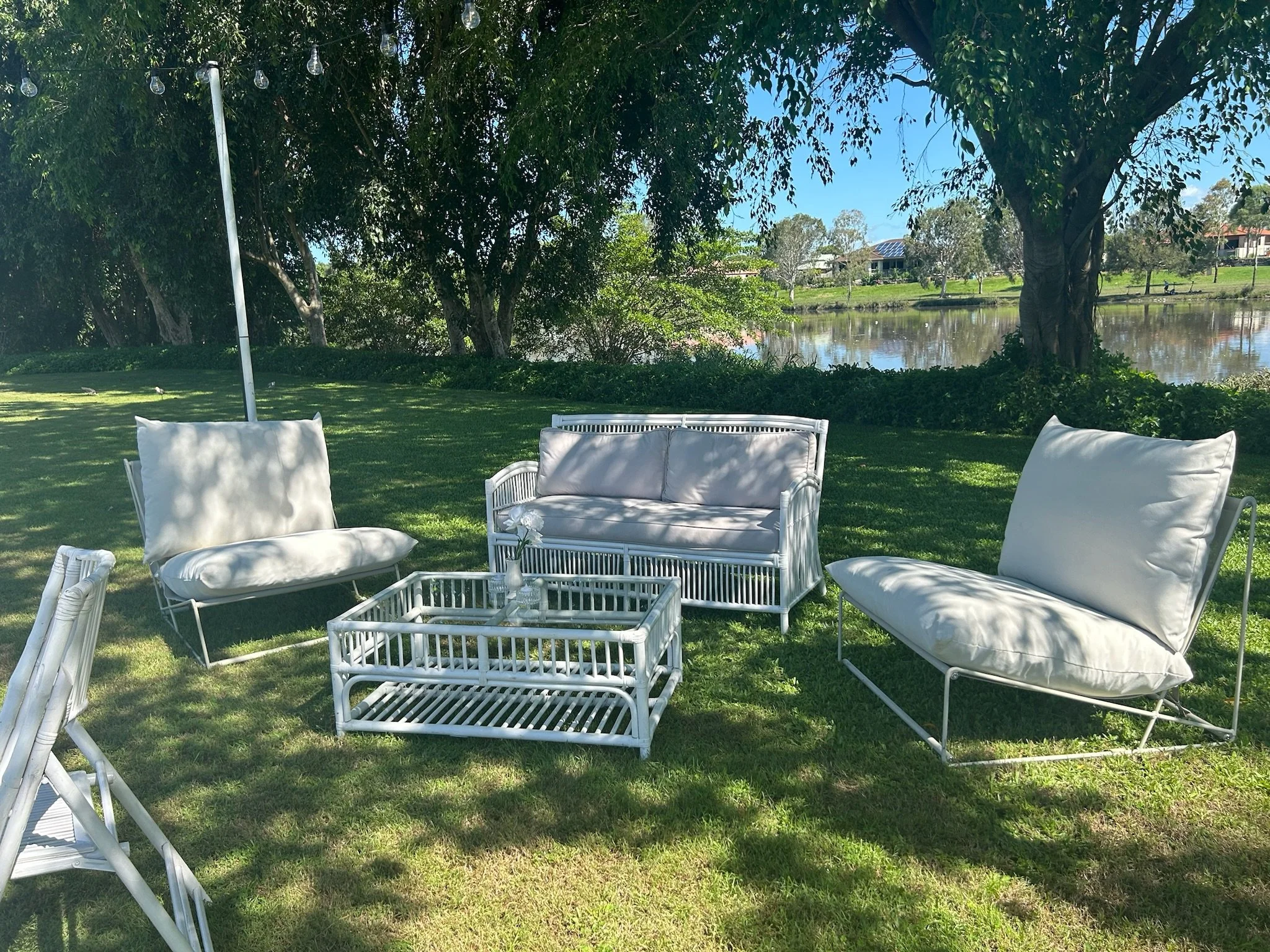 Outdoor patio furniture set with white cushioned chairs and a glass-topped table on a grassy lawn, near a lake with trees and houses in the background, under a partly sunny sky.