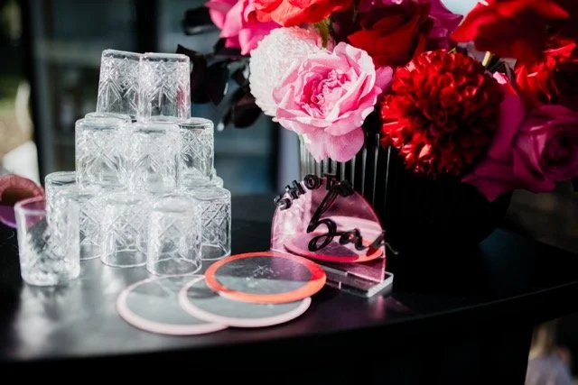 Stacked glassware, pink and red flowers in a black vase, and some jewelry and decorative items on a dark table.
