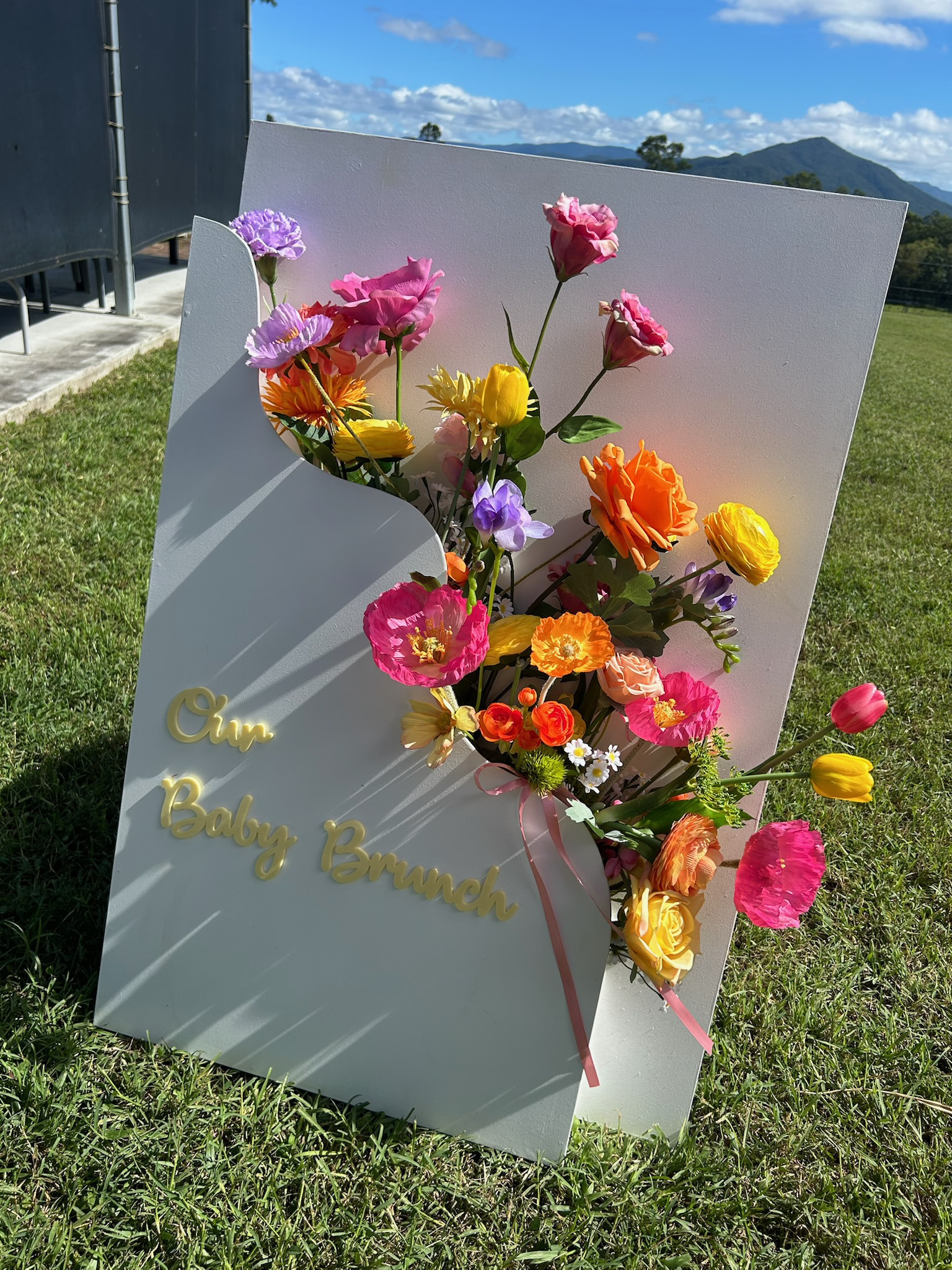 A white flower arrangement board with colorful flowers and the words 'Our Baby Brunch' written in light-colored cursive on it, outdoors on green grass with a mountain view and blue sky with clouds in the background.