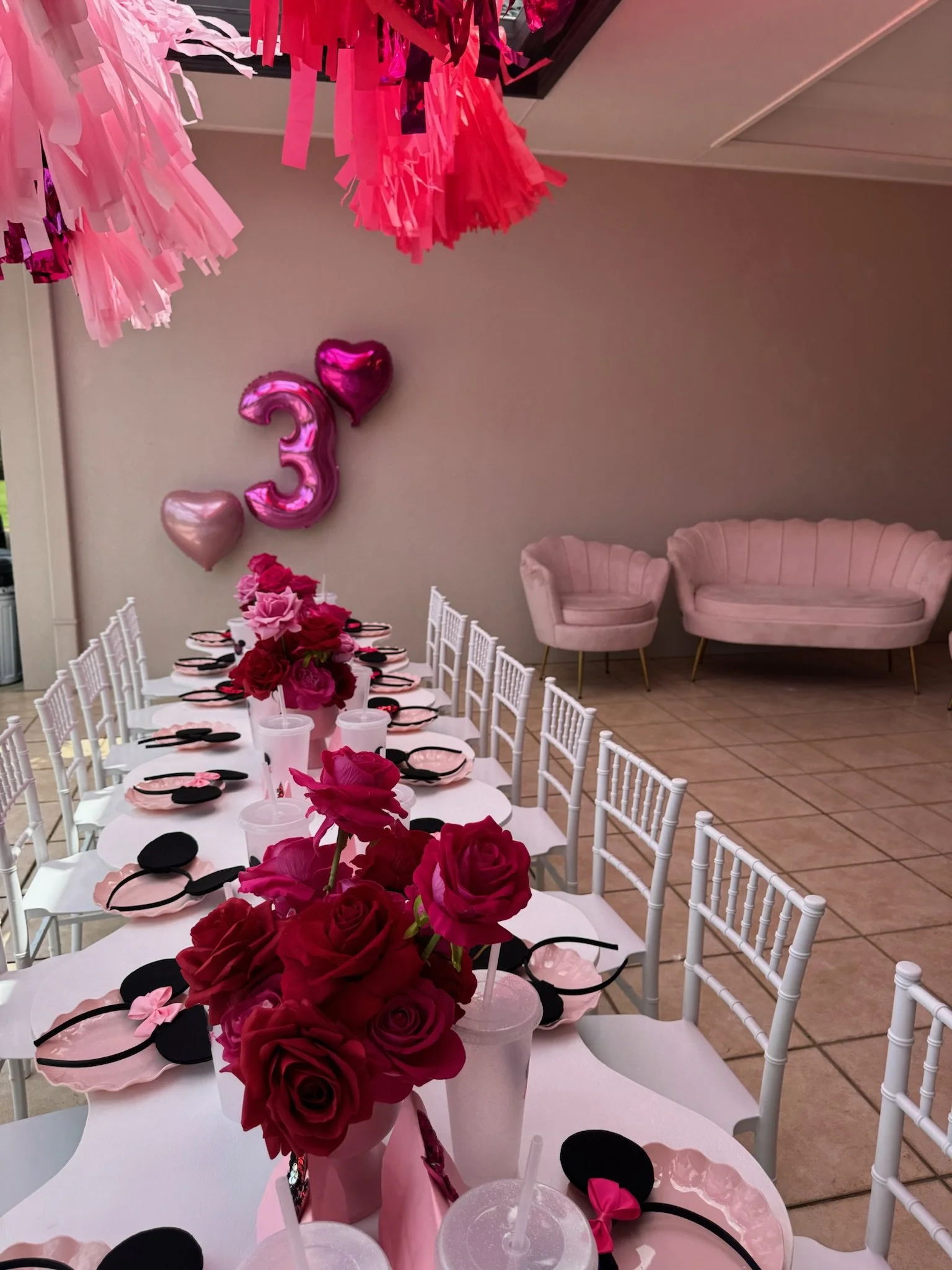 Decorations for a 3rd birthday party featuring pink, red, and black themed table settings with pink roses, balloons, and pink and black hats. Pink and black tissue paper tassels hanging from the ceiling. Pink plush sofas in the background.