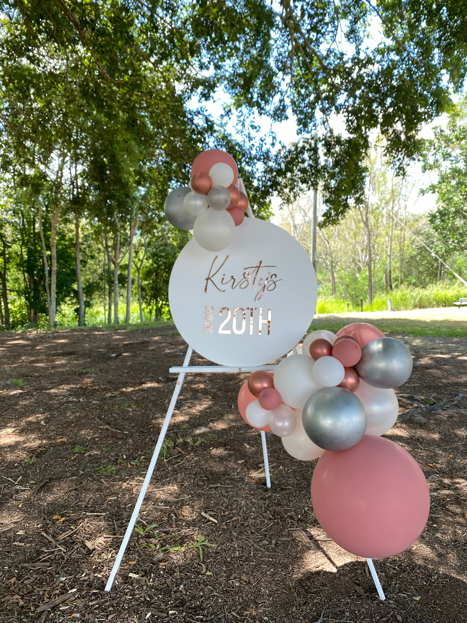 A decorative outdoor sign with balloons celebrating a 20th birthday, placed on a grassy area with trees and sunlight in the background.