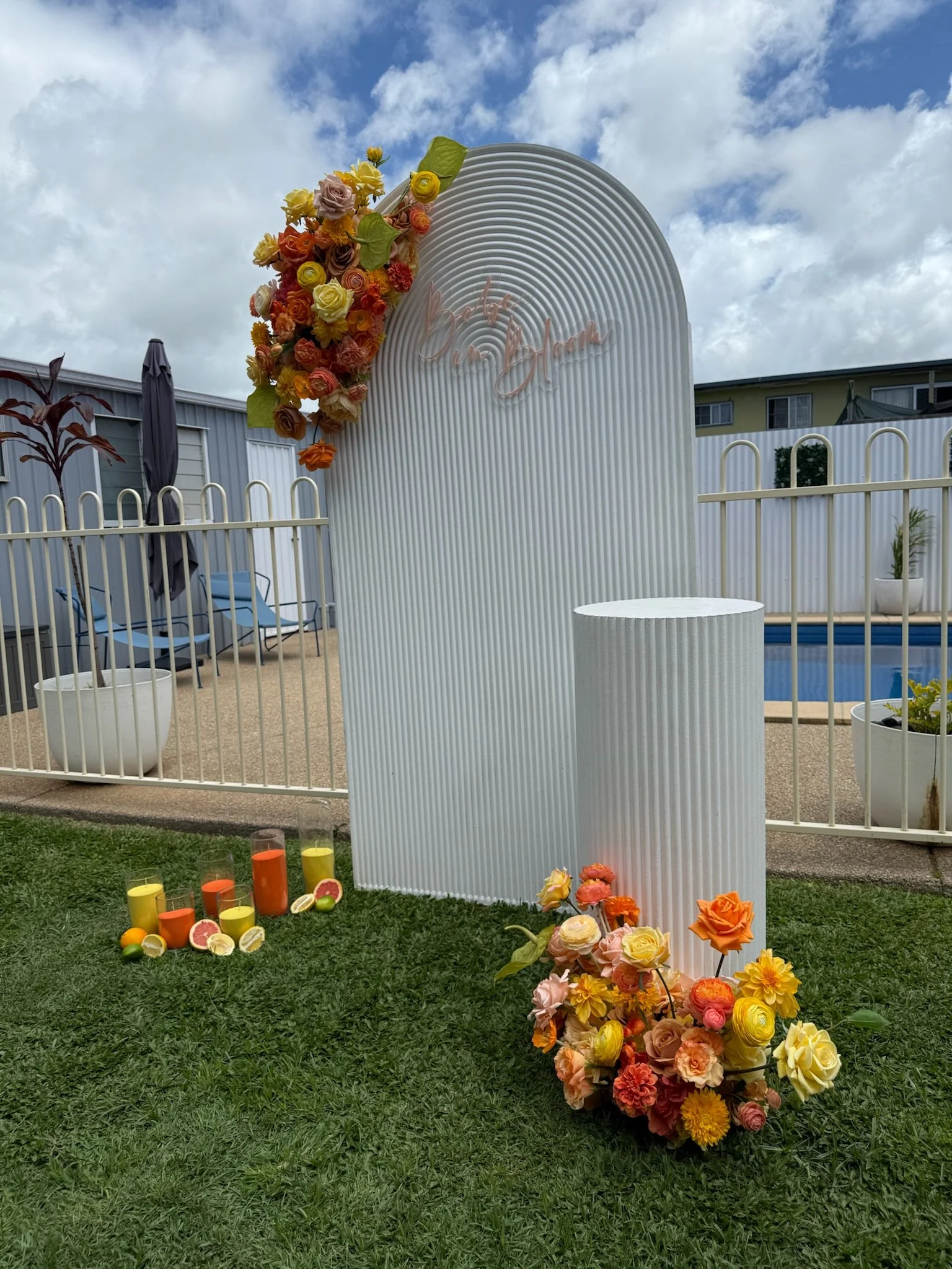 Decorative outdoor setup with floral arrangements, candles, and a white backdrop with pink script sign, near a pool and a backyard fence.