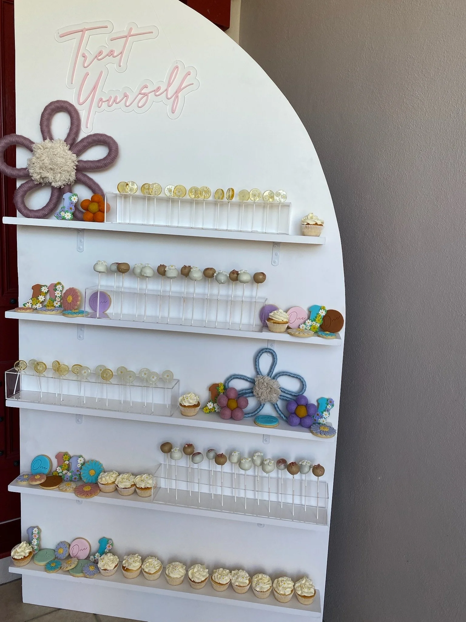 Decorative display board with cupcakes, cookies, and flower decorations, with a pink neon sign that says 'Treat Yourself' at the top.