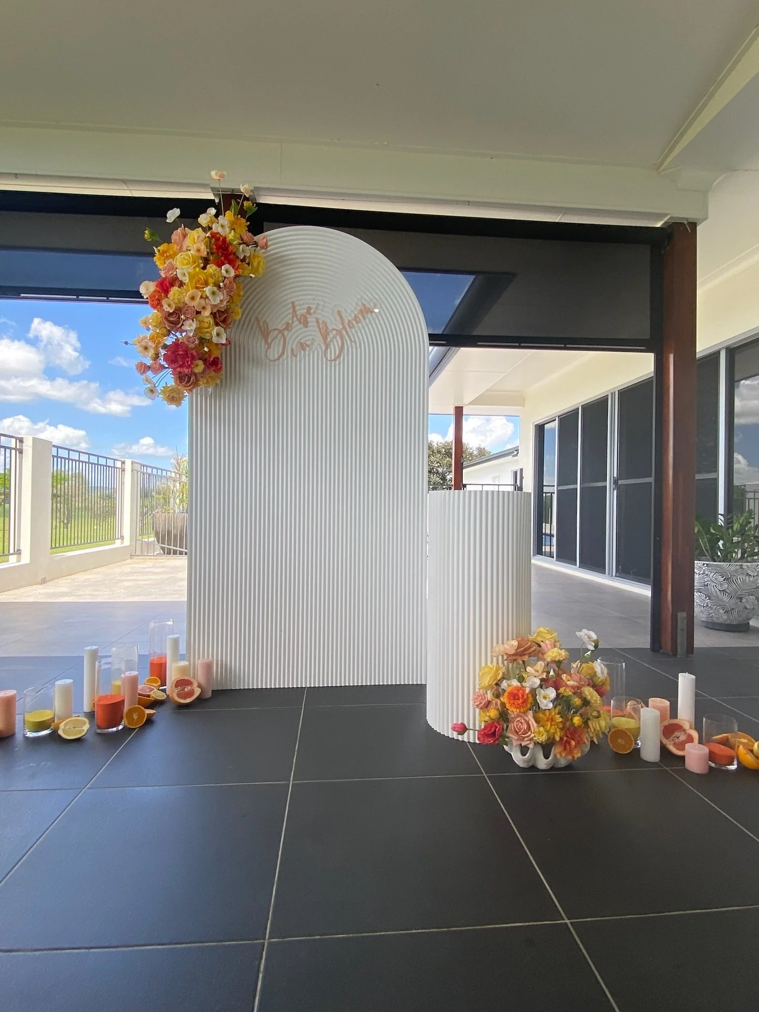 Decorative floral arrangement with candles and a sign with cursive writing on a white, curved backdrop on a covered patio with large windows, overlooking a blue sky and landscape.