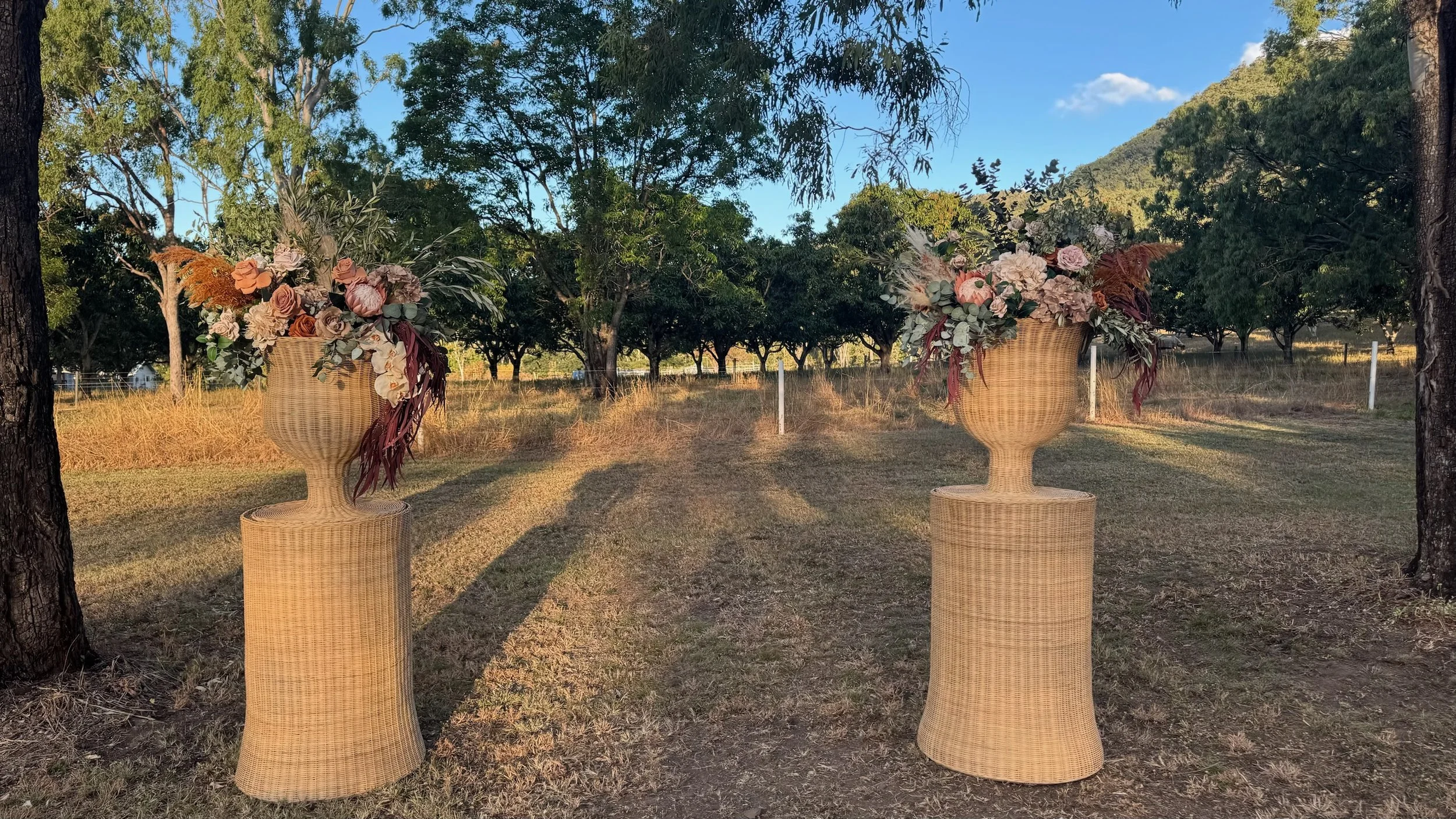 Two large wicker vases filled with dried flowers in shades of pink, cream, and brown, placed outdoor on grassy ground with trees and mountains in the background during sunset.