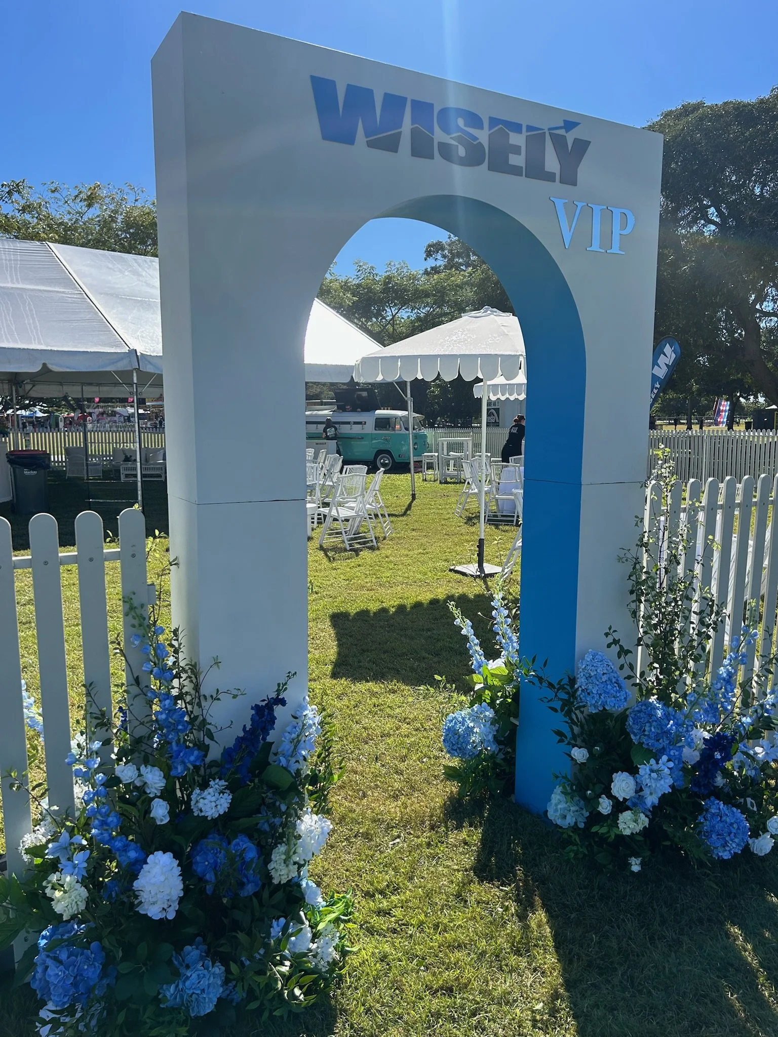 Entrance archway with the word 'WISLY' and 'VIP' above and decorated with white and blue flowers at its base. There are white outdoor chairs and tables inside a fenced outdoor area with tents and a vintage Volkswagen van in the background.