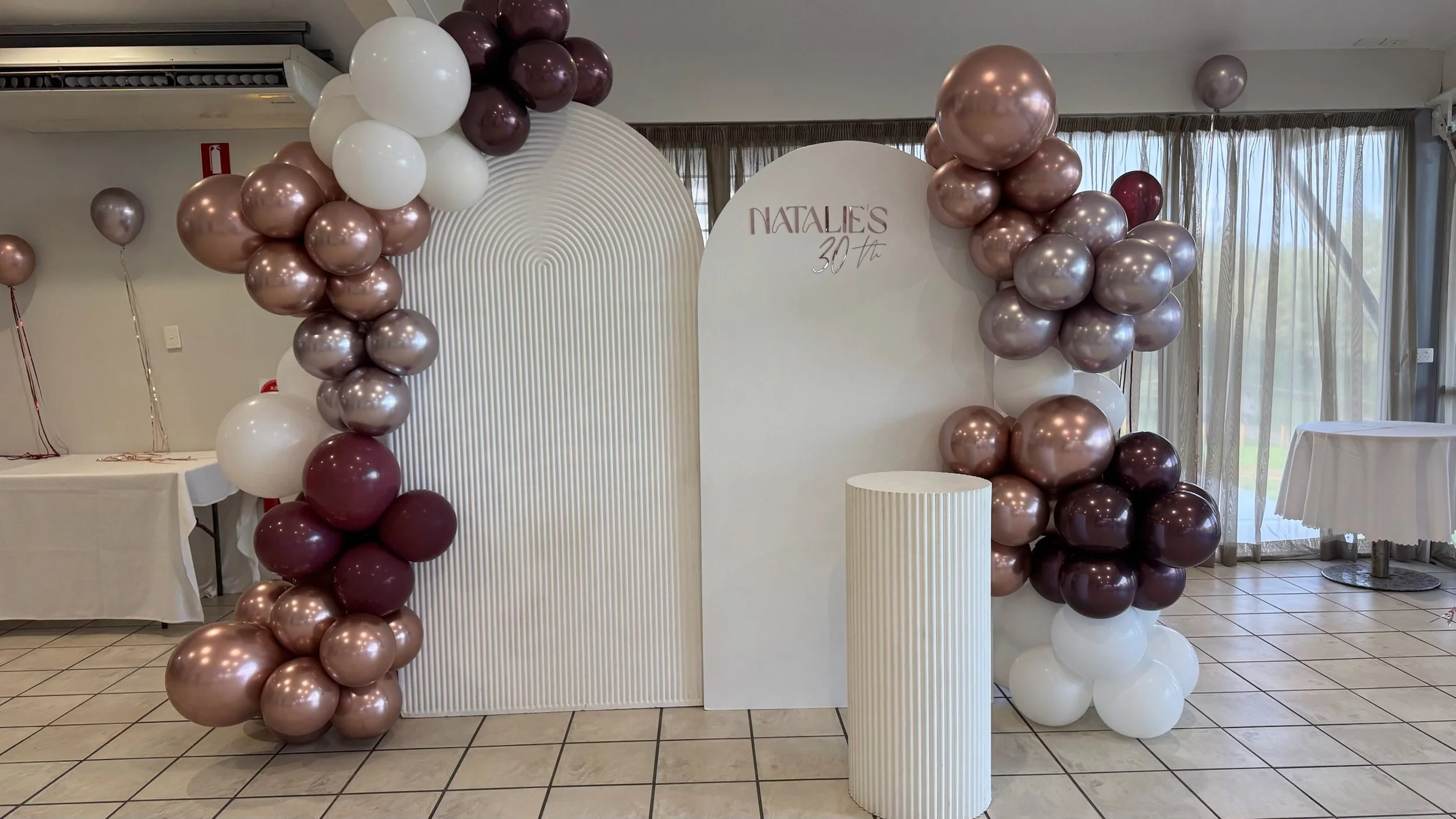 Decorative balloon arch with shades of rose gold, white, and burgundy balloons at a birthday celebration, with a backdrop reading 'NATALIE'S 30th'.
