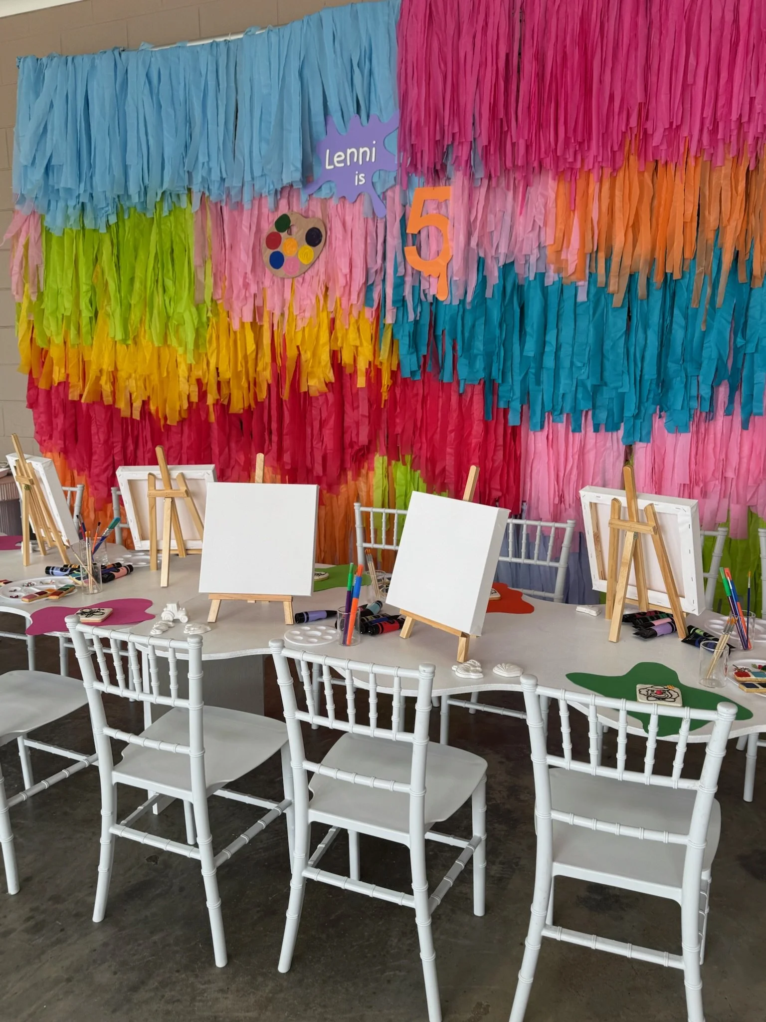 Colorful party or art studio setup with a large vibrant backdrop made of multicolored tissue paper fringe, featuring signs that say "Lenni is 5" and a paint palette illustration, with empty canvases, paint supplies, and white chairs arranged on a tab