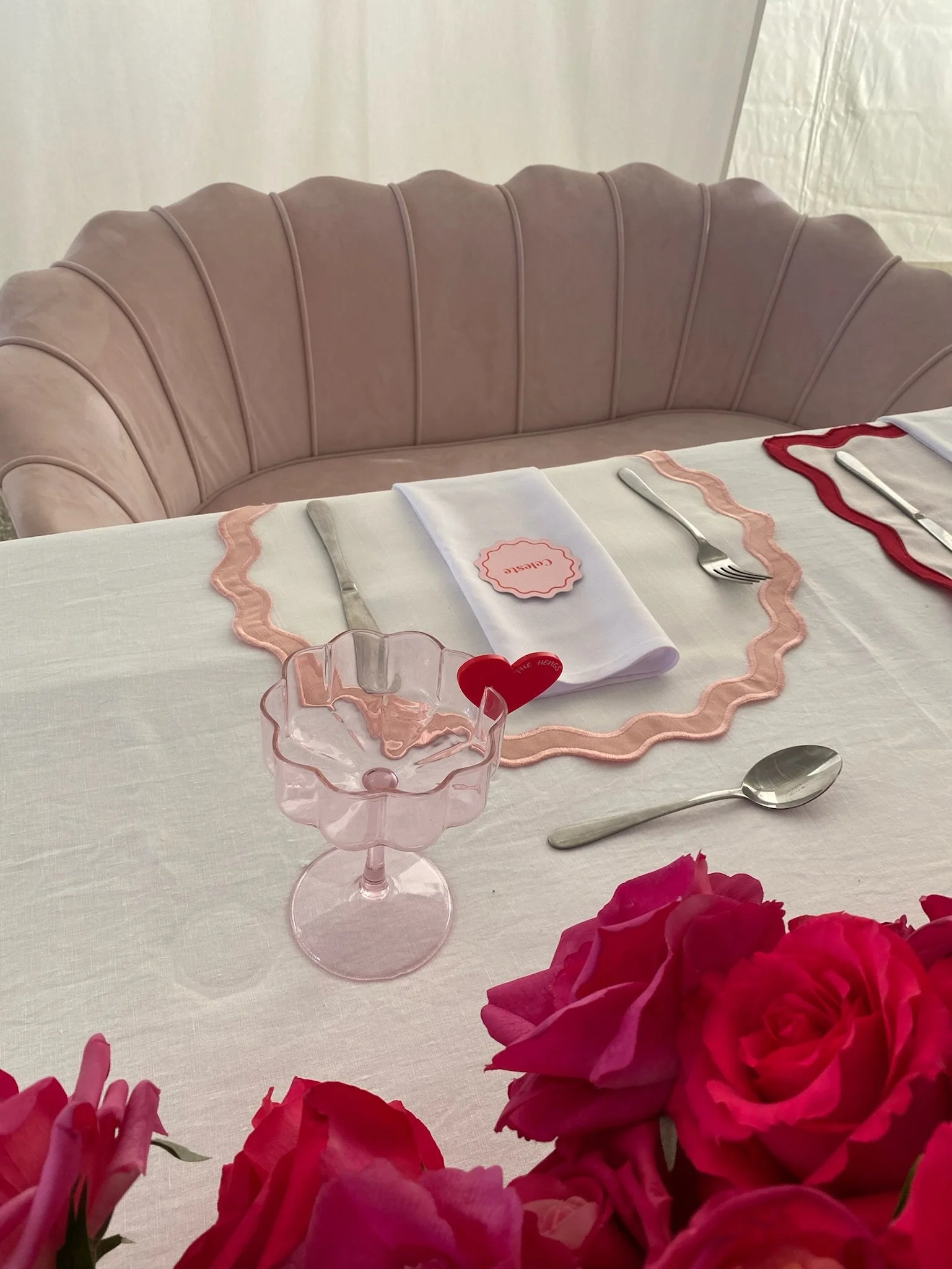 A pink velvet sofa behind a decorated dining table with pink roses, a pink glass dessert cup, a white cloth napkin with a pink scalloped edge, and a small red heart-shaped decoration.