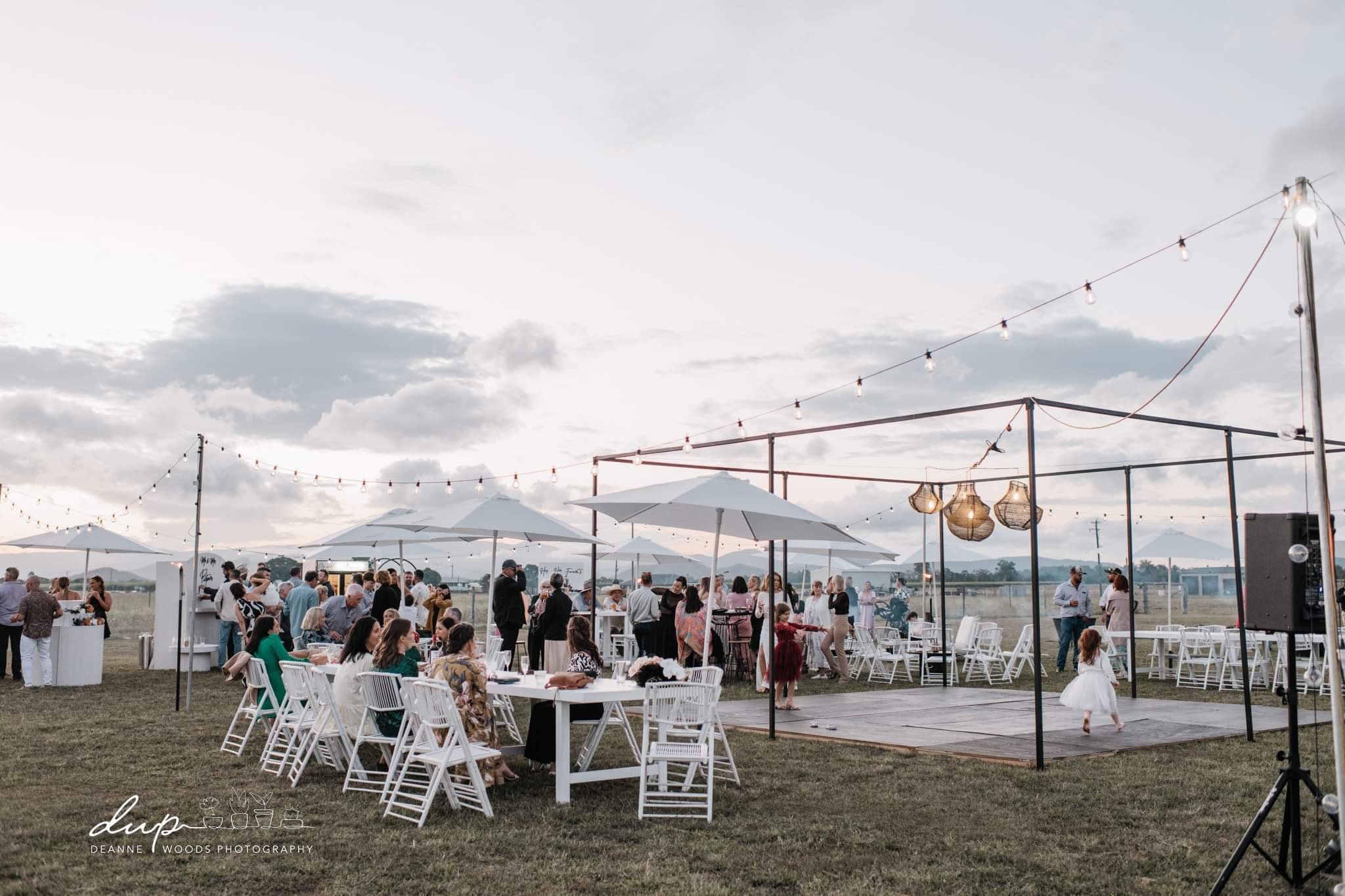 Outdoor wedding reception with guests, tables, umbrellas, string lights, a dance floor, and a cloudy sky.