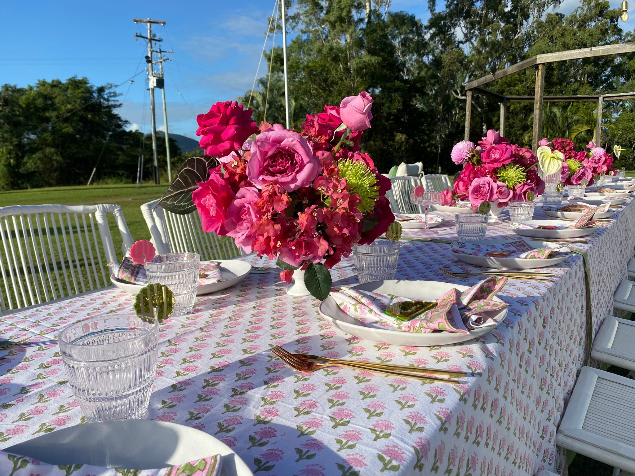 Long outdoor dining table decorated with pink flower arrangements, gold utensils, patterned napkins, and glassware, set-up for a celebration in a grassy area with trees and cloudy sky in the background.