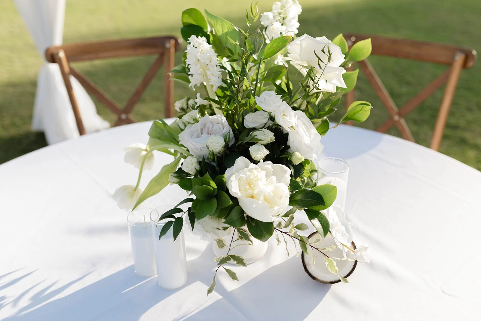 White floral arrangement on a round white table with wooden chairs in the background, outdoors