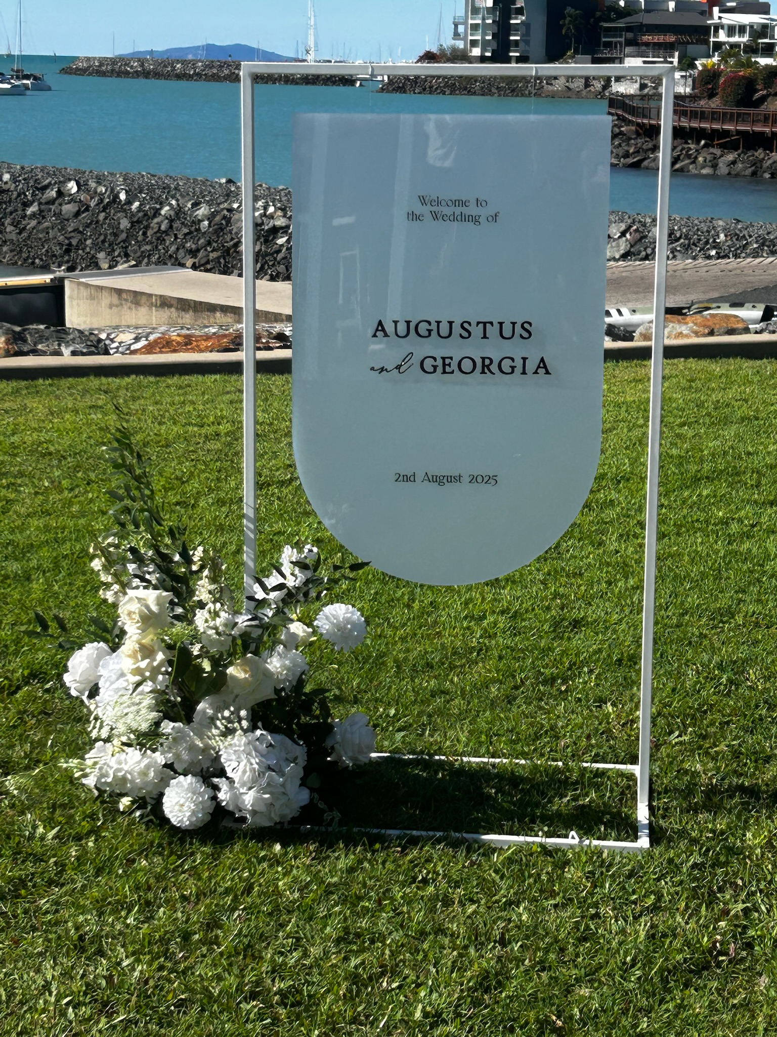 A wedding sign on a grassy area by the water, with a bouquet of white flowers at the base, announcing the wedding of August and Georgia on August 2, 2025.