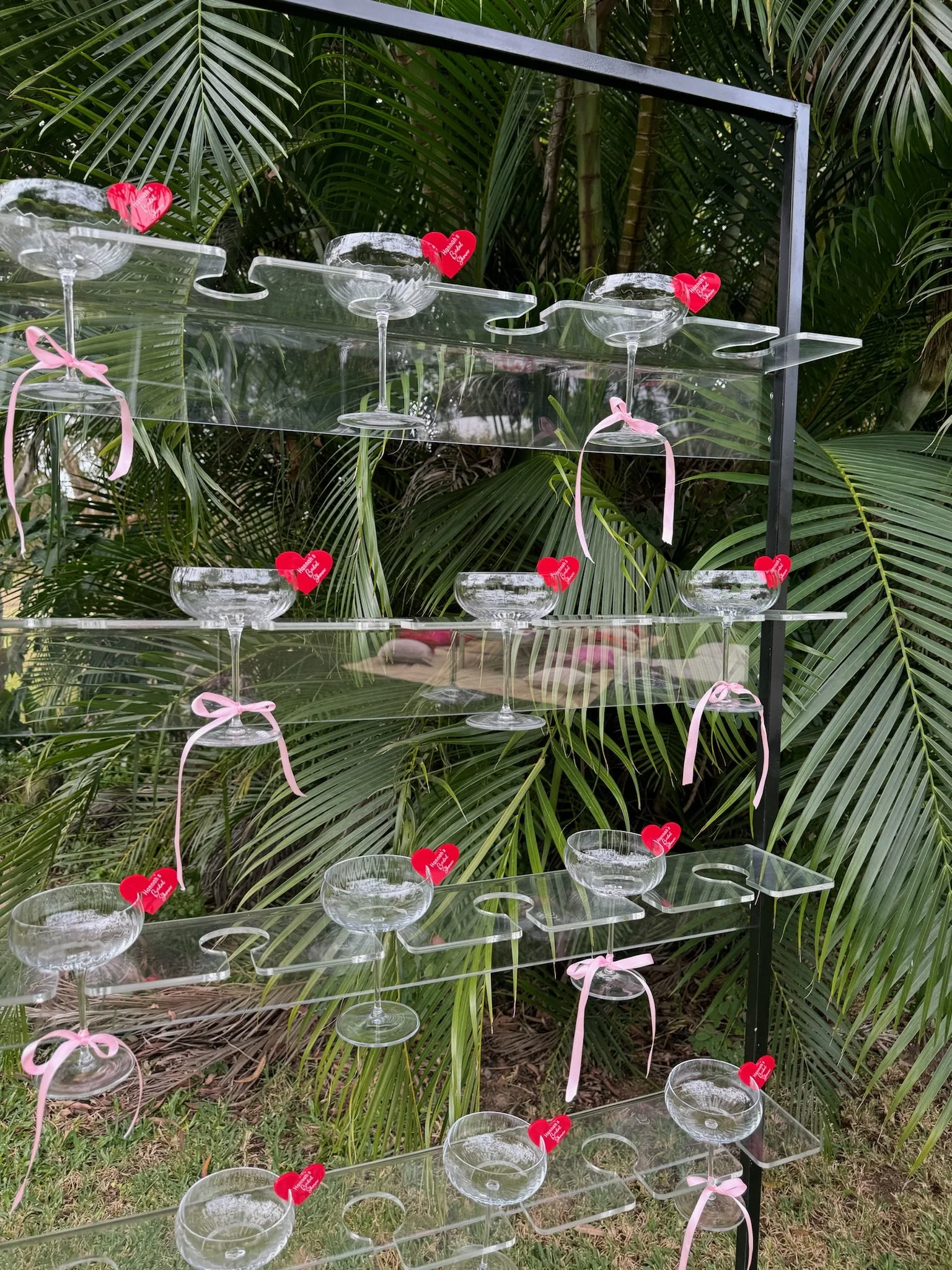 A black metal and acrylic shelving unit outdoors, decorated with small red heart-shaped tags, pink ribbons, and clear glass champagne coupes, with large green palm leaves in the background.