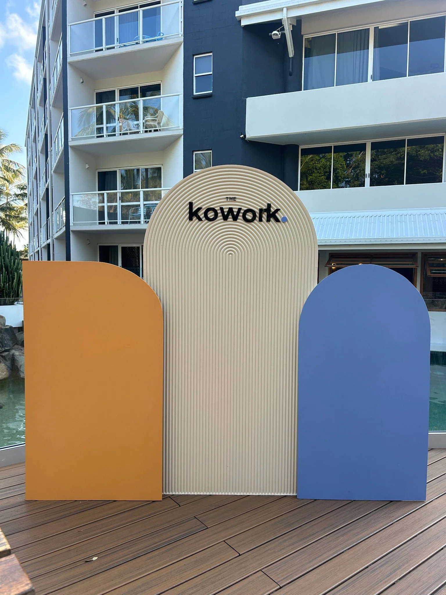 A decorative sign reads 'The Kowork' with colorful curved panels in yellow and blue in front of a modern multi-story apartment building with balconies and glass railings.