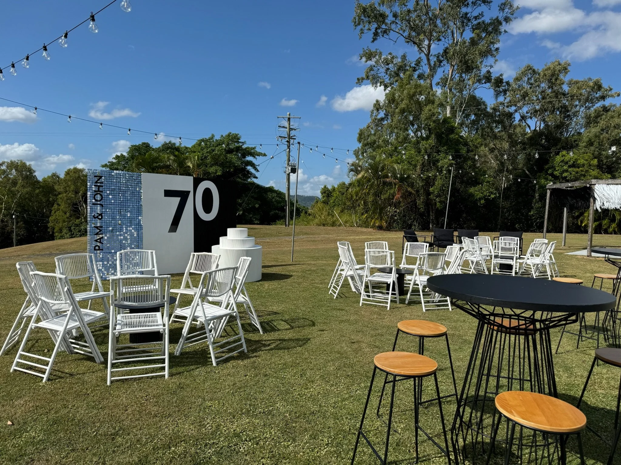Outdoor event setup with white chairs, black tables, and a large sign reading '70'. There are string lights hanging above, trees in the background, and a clear blue sky.